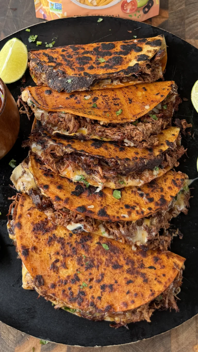 Stack of crispy birria tacos with melted cheese and shredded beef on a black platter with lime wedges