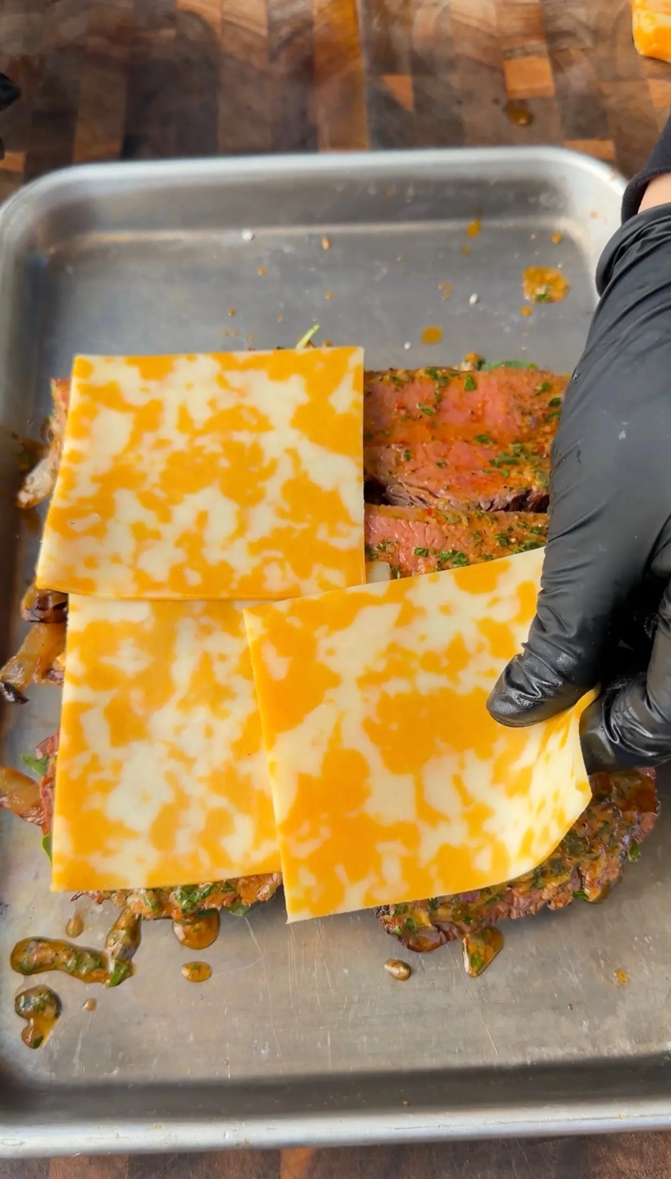 Gloved hand placing colby jack cheese slices over seasoned tri-tip sliders with cowboy butter sauce in a metal tray