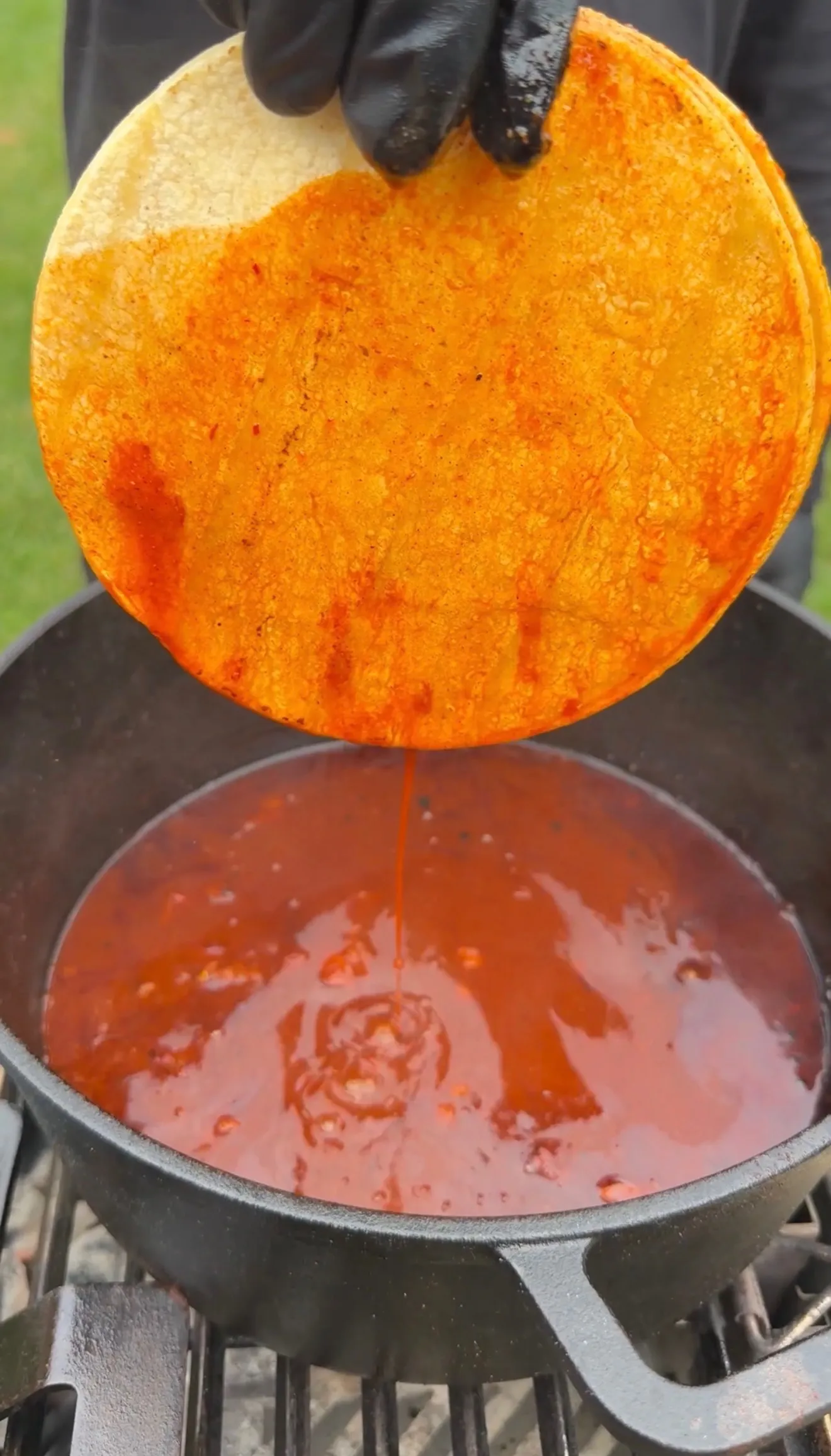 Gloved hand dipping a corn tortilla into rich red birria consommé in a cast iron pot on a grill