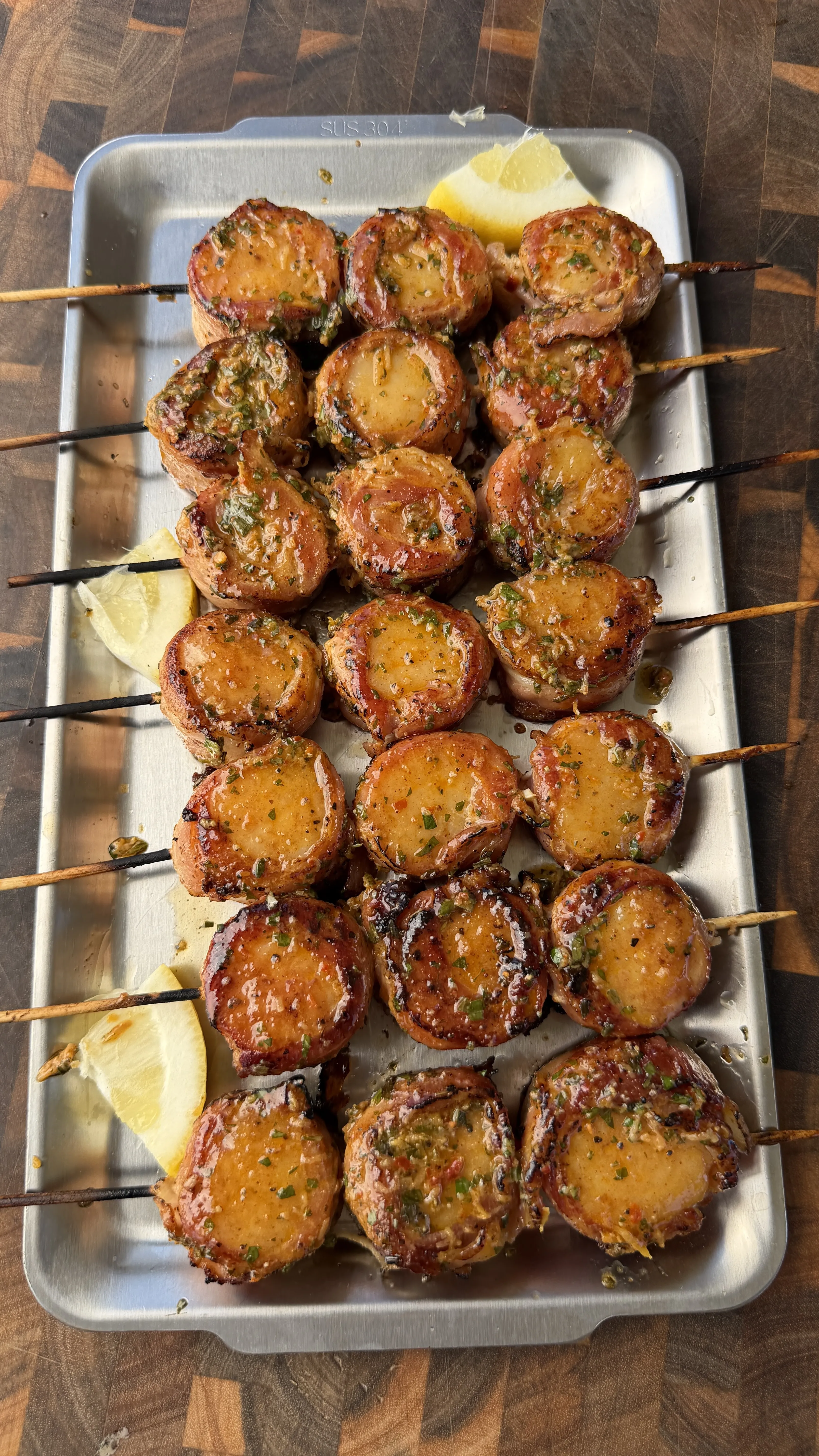 Grilled bacon-wrapped scallop skewers glazed with cowboy butter on a metal tray with lemon wedges