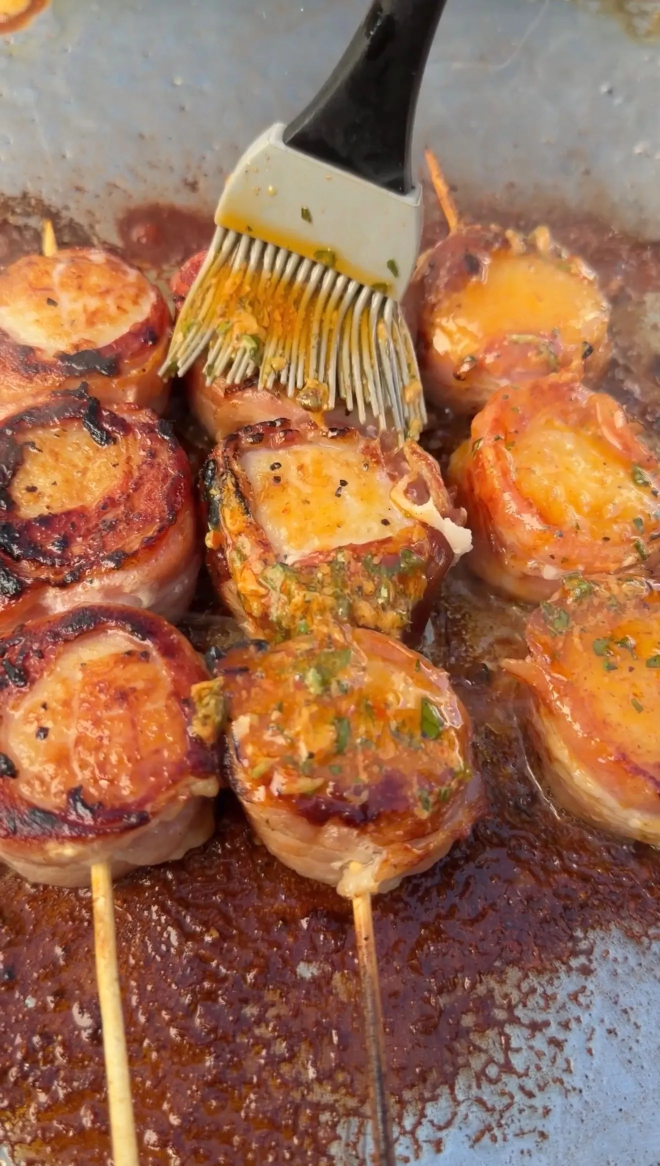 Bacon-wrapped scallops on skewers being basted with cowboy butter herb sauce on a sizzling pan