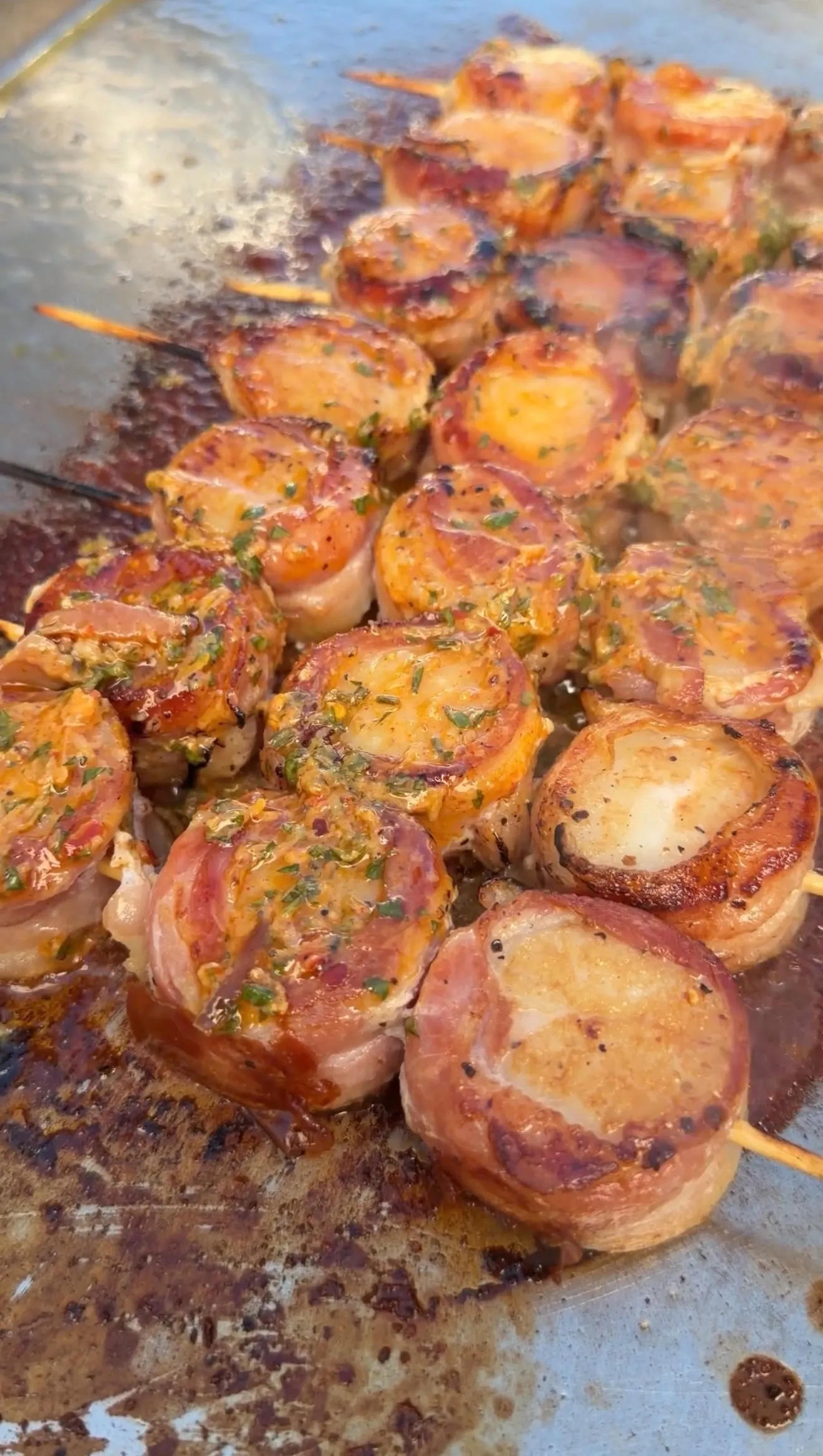 Bacon-wrapped scallops on skewers sizzling on a flat-top griddle, glazed with cowboy butter and fresh herbs