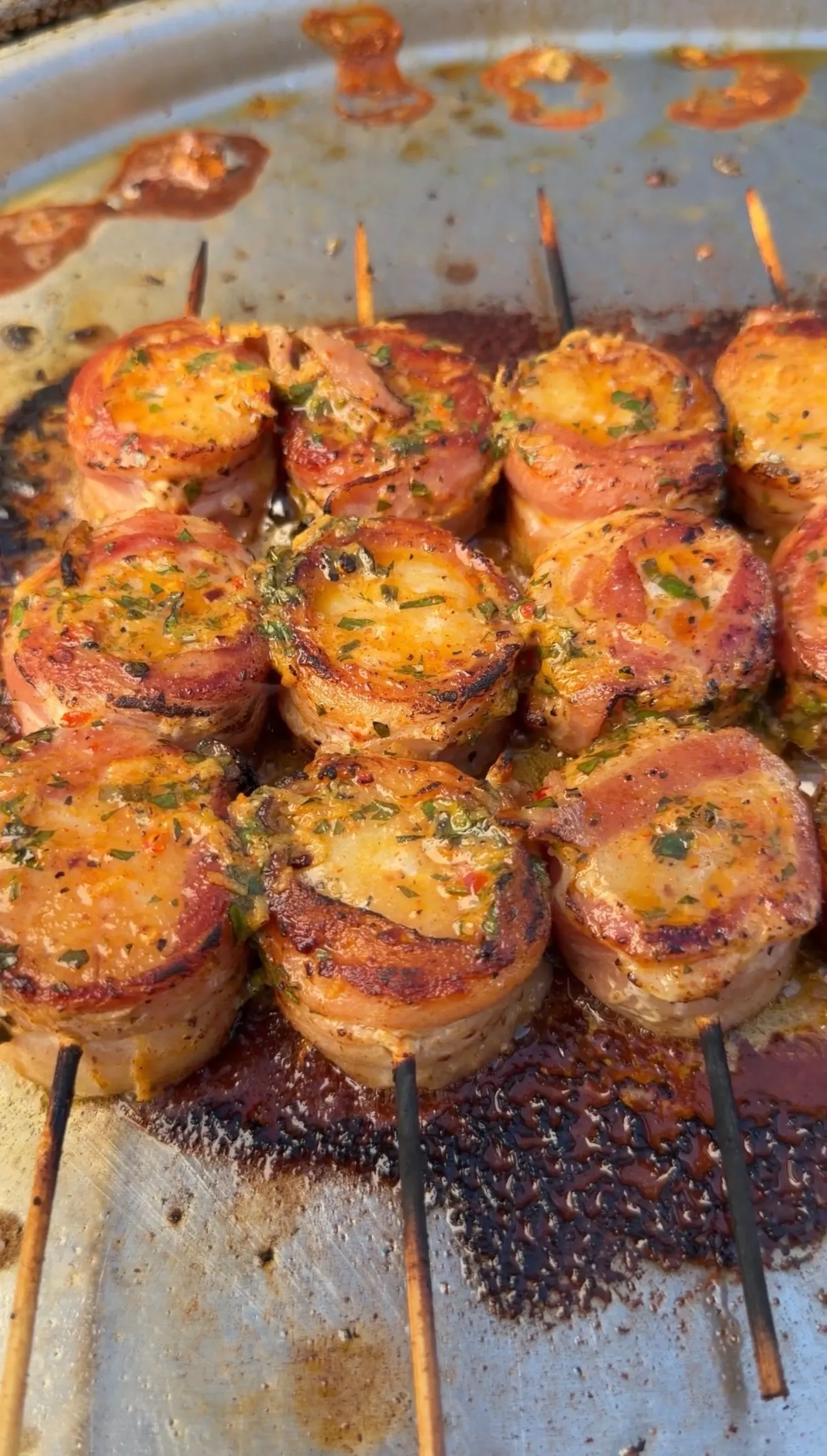 Bacon-wrapped scallops on skewers coated in cowboy butter herb sauce, sizzling on a round grill pan