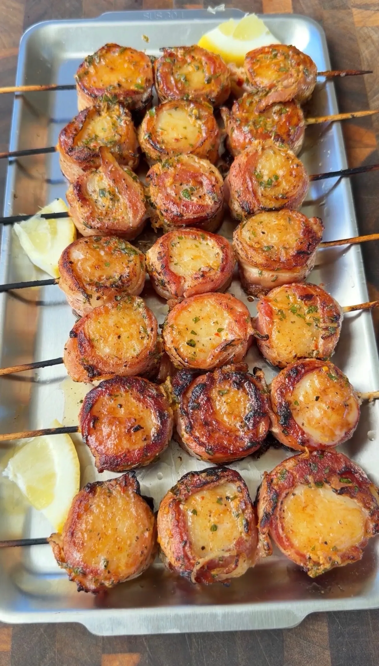 Bacon-wrapped scallops on skewers with cowboy butter seasoning, served on a metal tray with lemon wedges