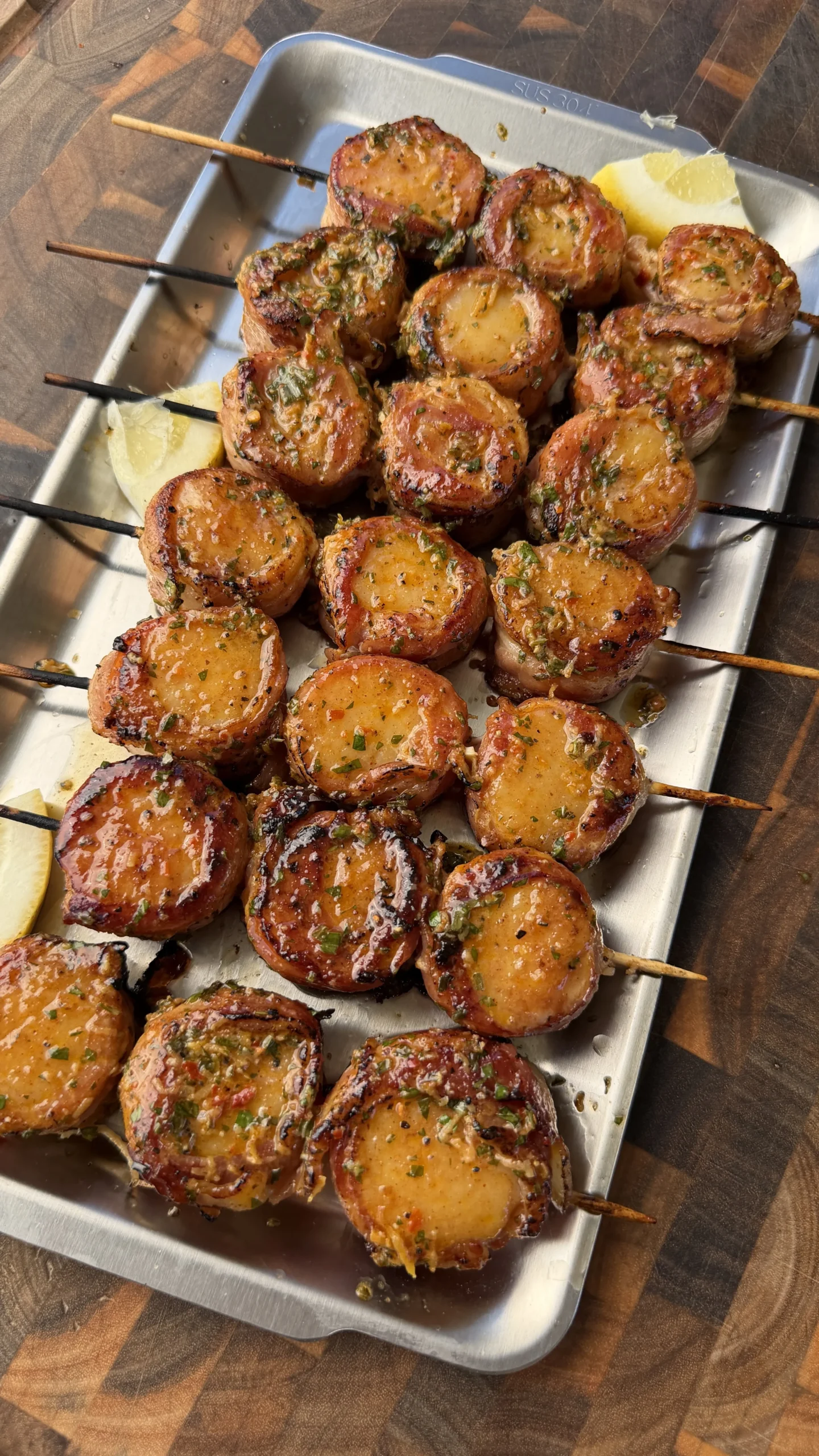 Grilled bacon-wrapped scallops on skewers glazed with cowboy butter, served on a metal tray with lemon wedges