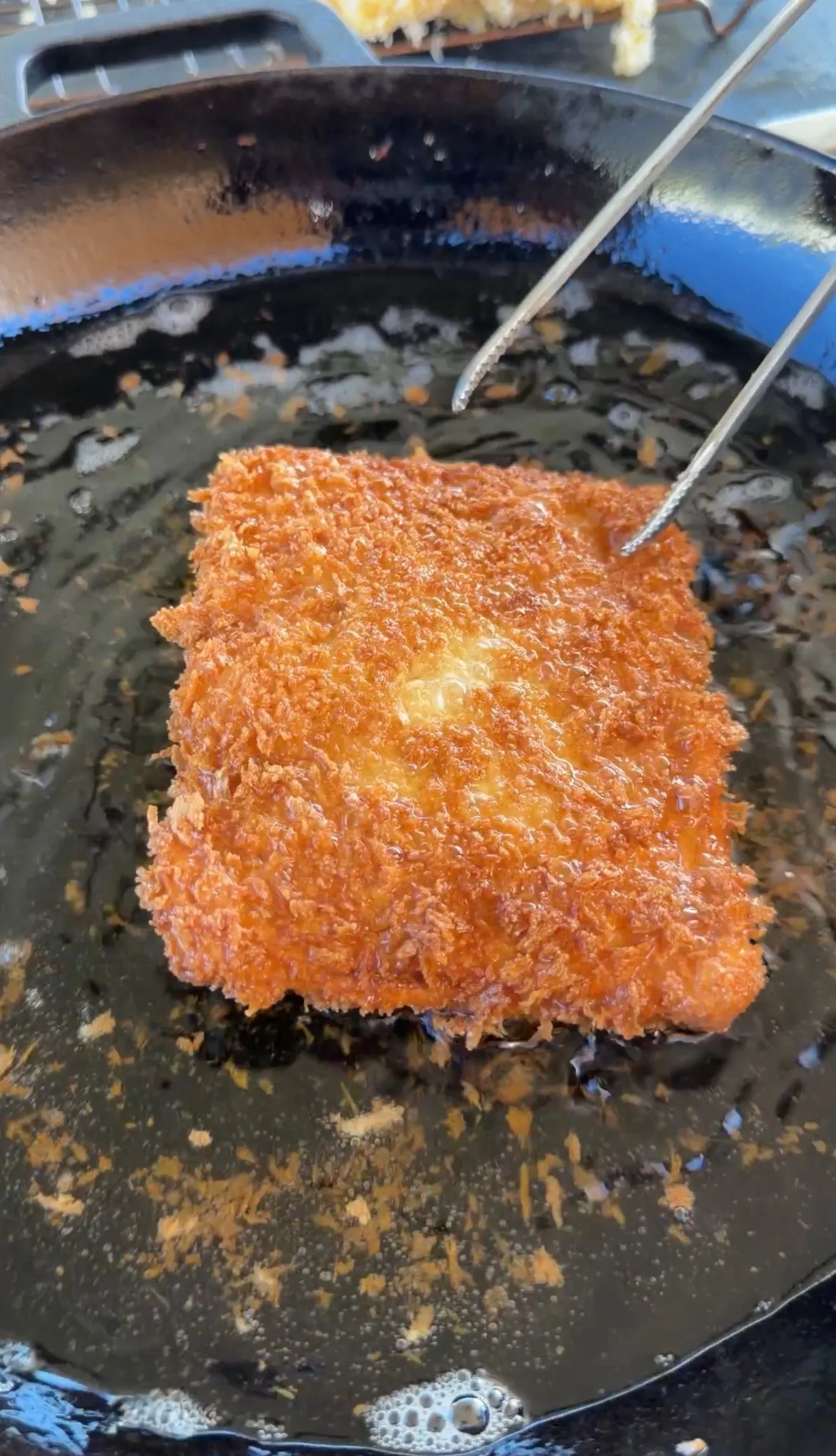 Golden-brown crispy breaded cheesesteak stuffed pocket frying in a cast iron skillet with tongs