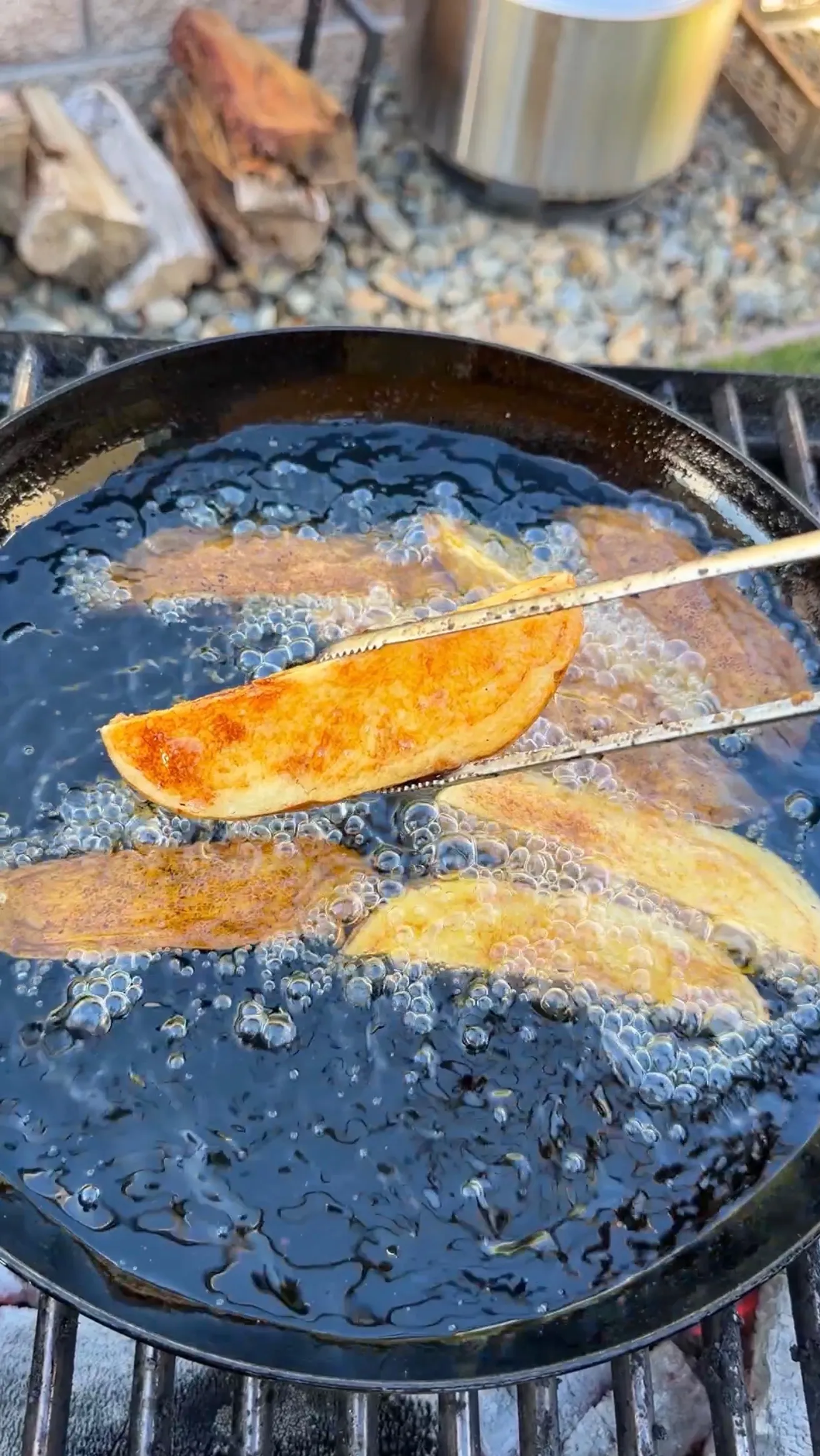 Golden crispy potato wedges deep frying in bubbling hot oil in a large cast iron skillet over outdoor grill