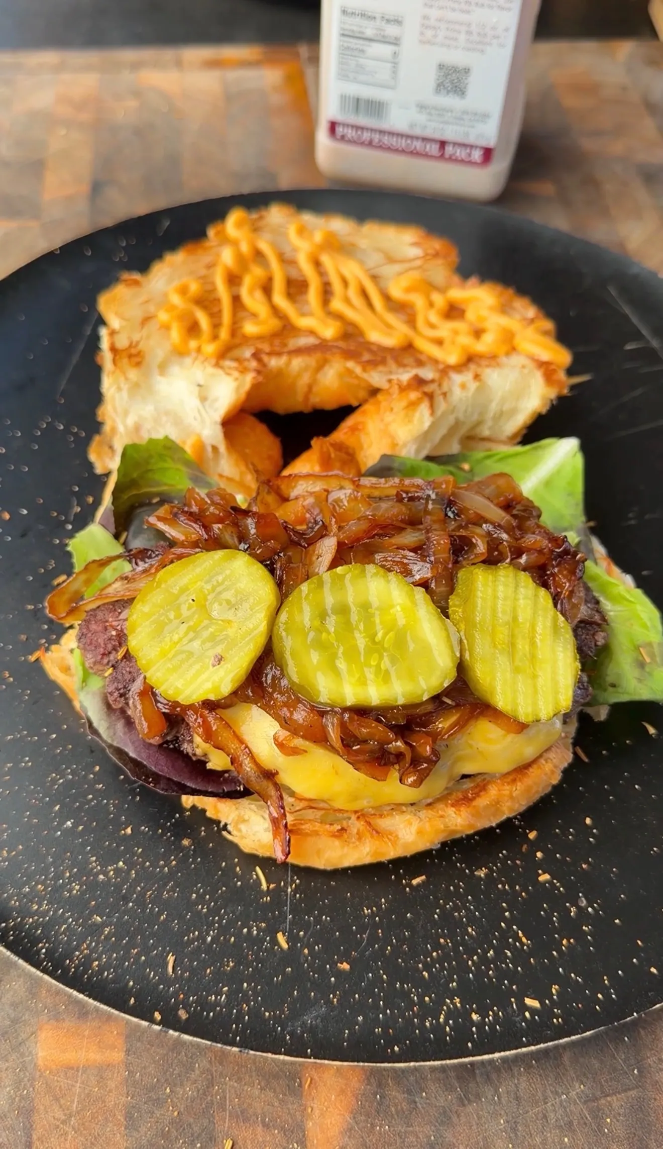 Open-face croissant burger topped with pickles, caramelized onions, melted cheese, and lettuce on a black plate