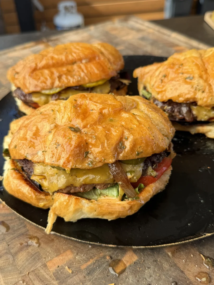 Juicy croissant burgers with melted cheese, pickles, tomato, and caramelized onions served on a black plate