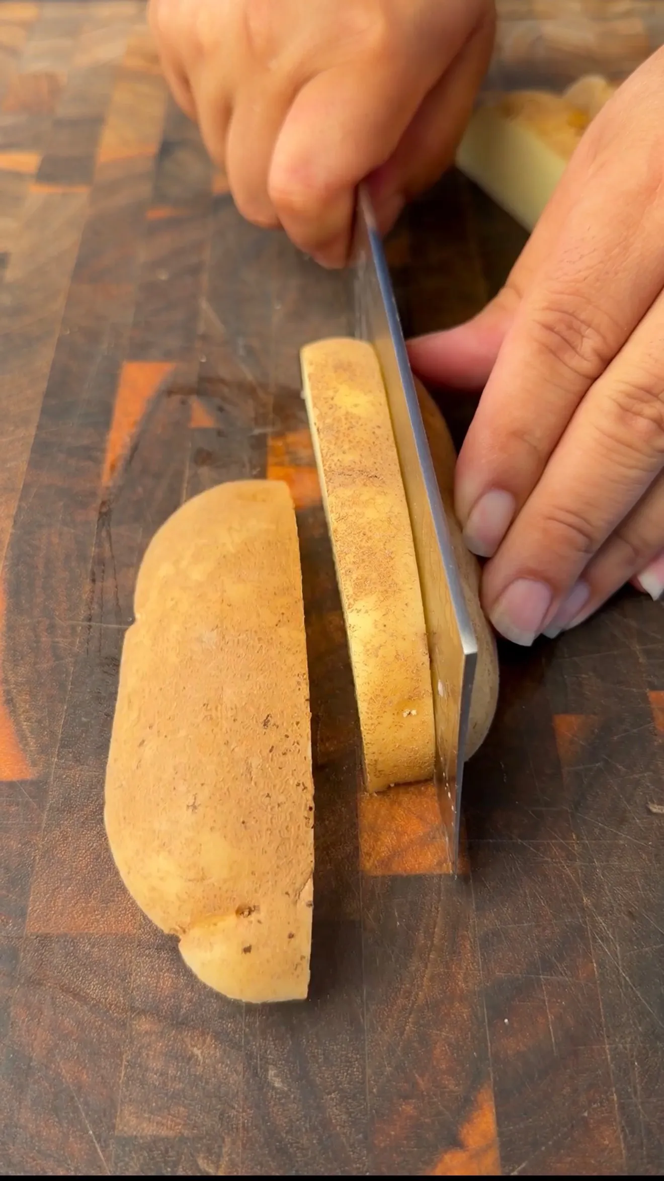 Hands slicing a russet potato into thick wedges with a chef's knife on a wooden cutting board