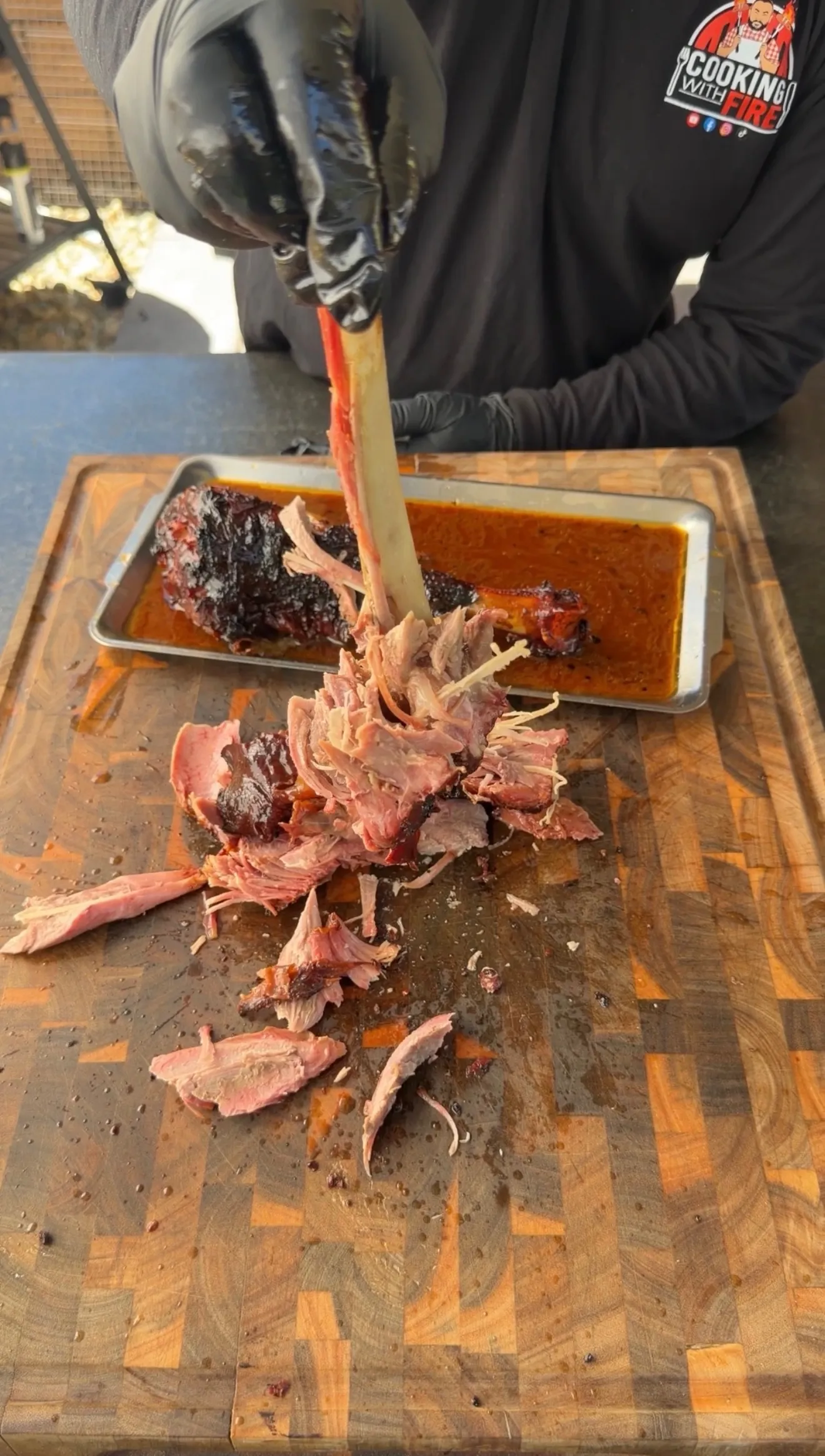 Gloved hands pulling apart a fall-apart smoked turkey leg on a wooden cutting board with rich sauce in background