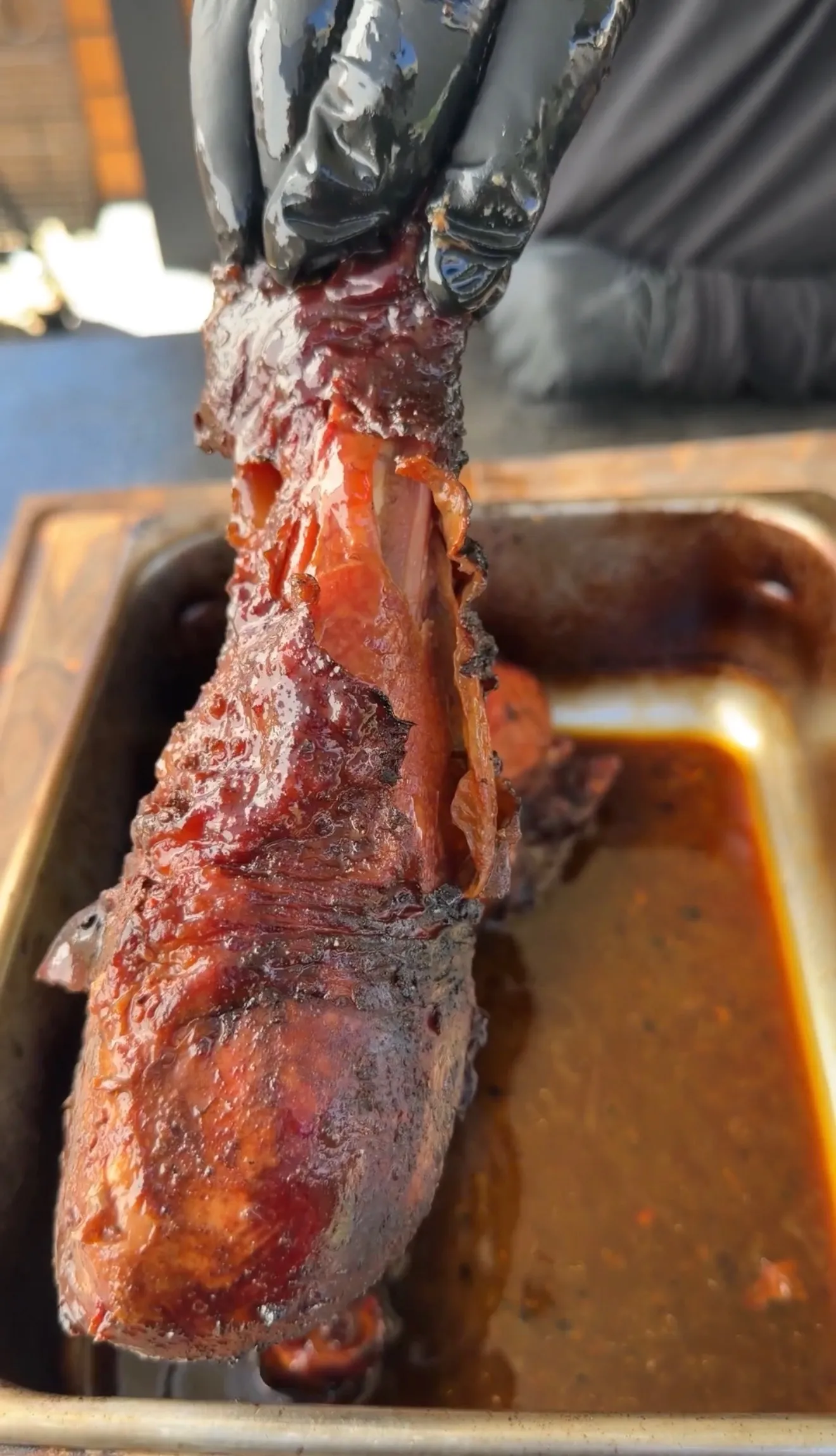 Glazed smoked turkey leg held up with black gloves above a pan of rich barbecue drippings