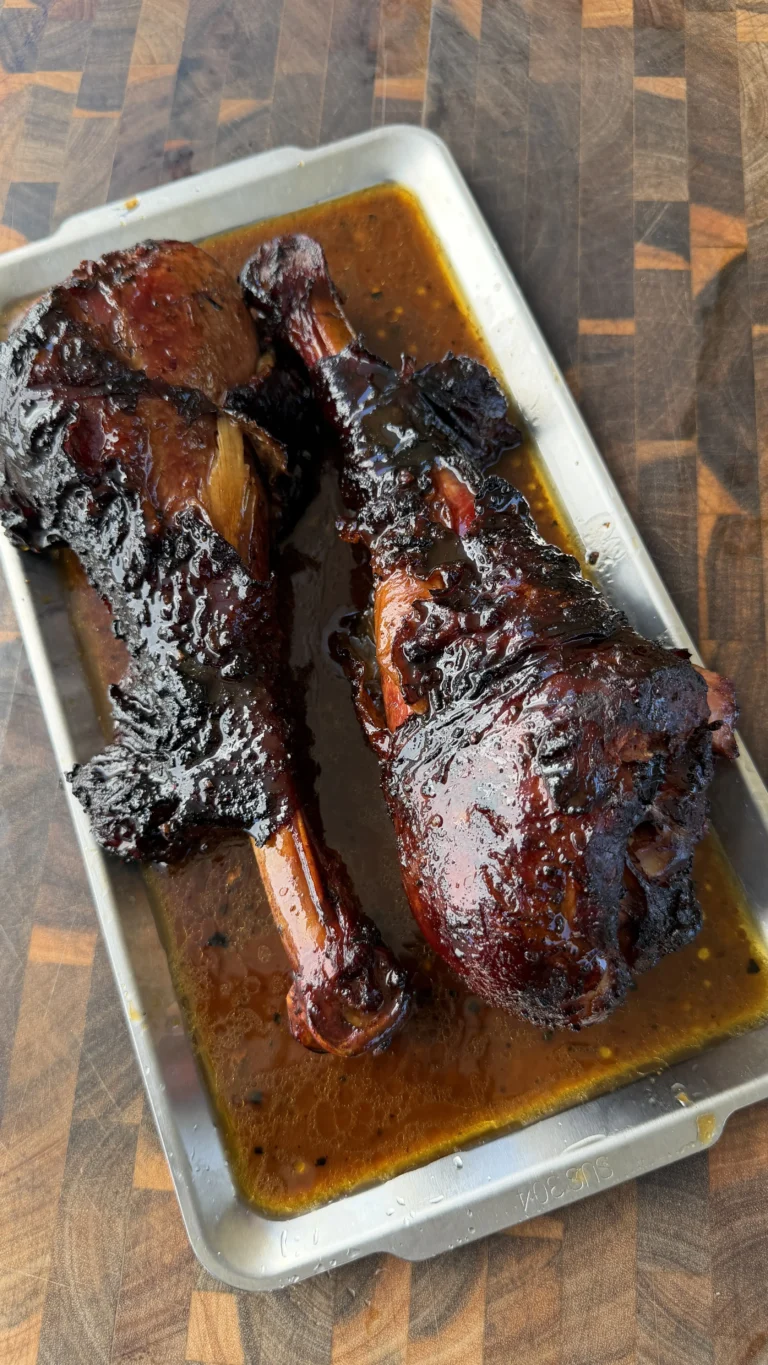 Two glazed smoked turkey legs resting in a metal pan with rich BBQ drippings on a wooden cutting board