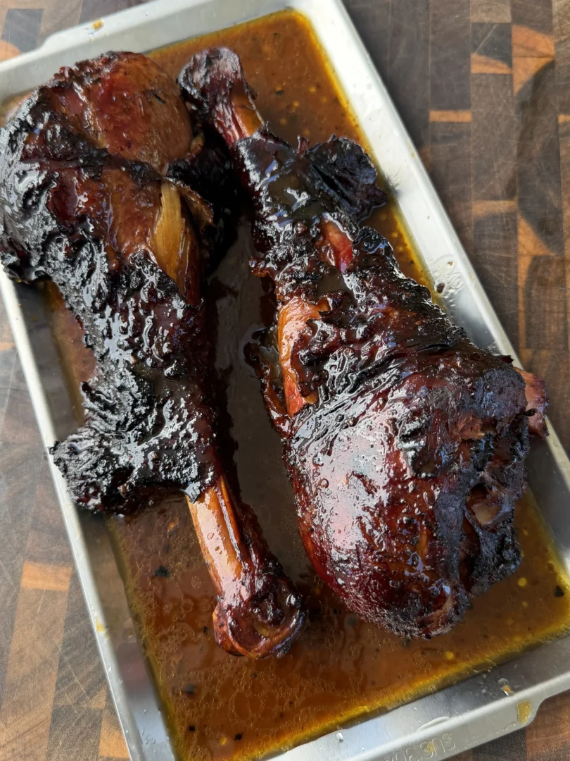 Two glazed smoked turkey legs resting in a metal pan with rich BBQ drippings on a wooden cutting board