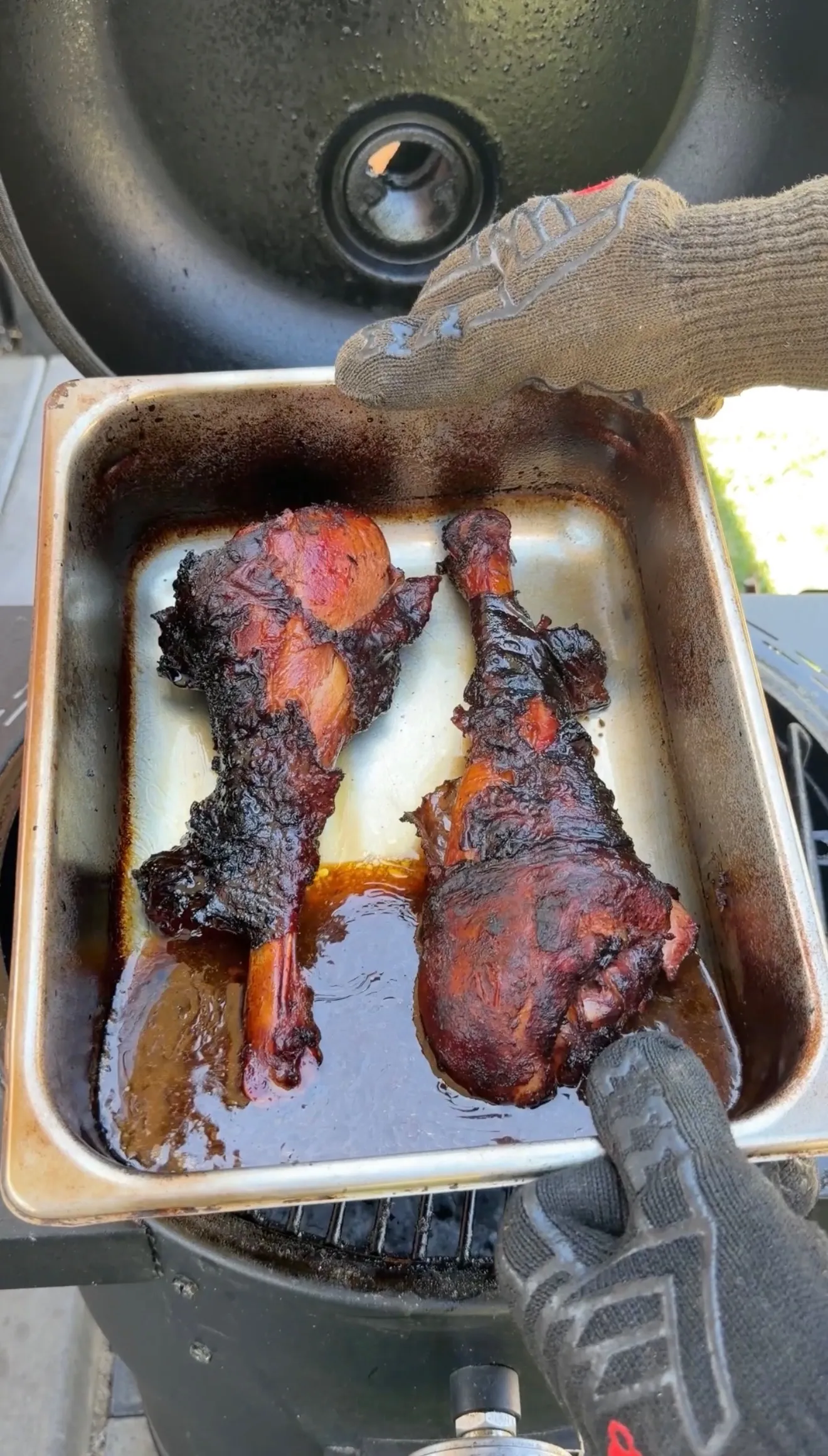 Two deeply smoked turkey legs in a caramelized drippings pan, held with BBQ gloves over a smoker