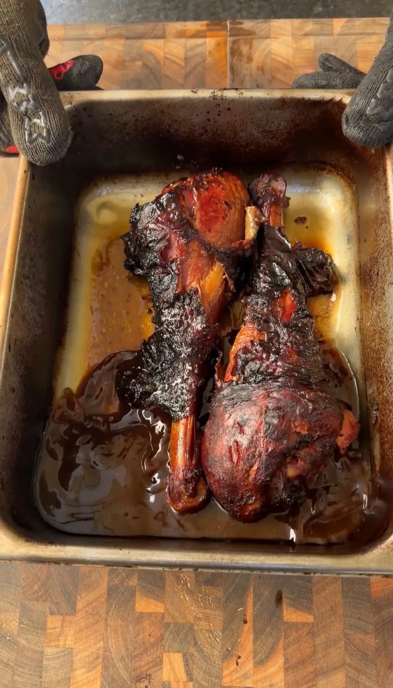 Two deeply smoked turkey legs resting in a roasting pan with rich dark juices, held by heat-resistant gloves