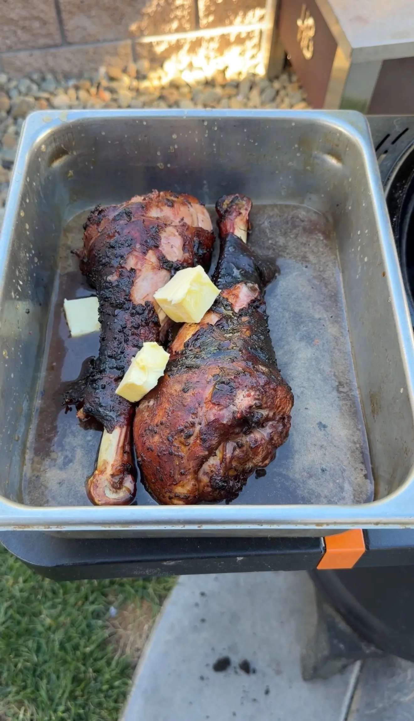 Two deeply smoked turkey legs in a steel pan topped with melting butter pats, resting in juices