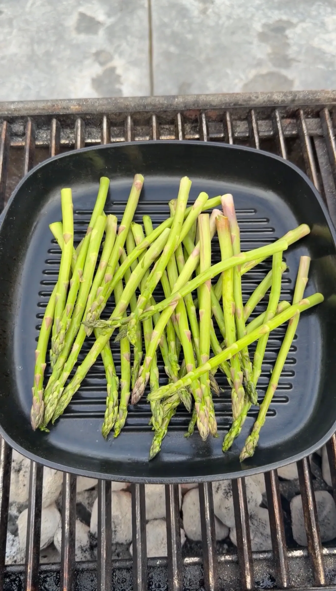 Fresh green asparagus spears arranged on a black ridged grill pan over charcoal grill grates