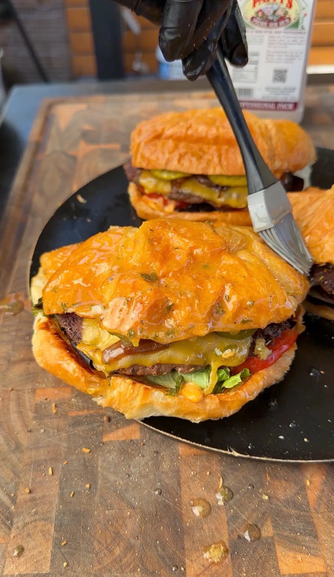 A double croissant burger being brushed with glaze, loaded with cheese, lettuce, tomato, and beef patties on a black plate