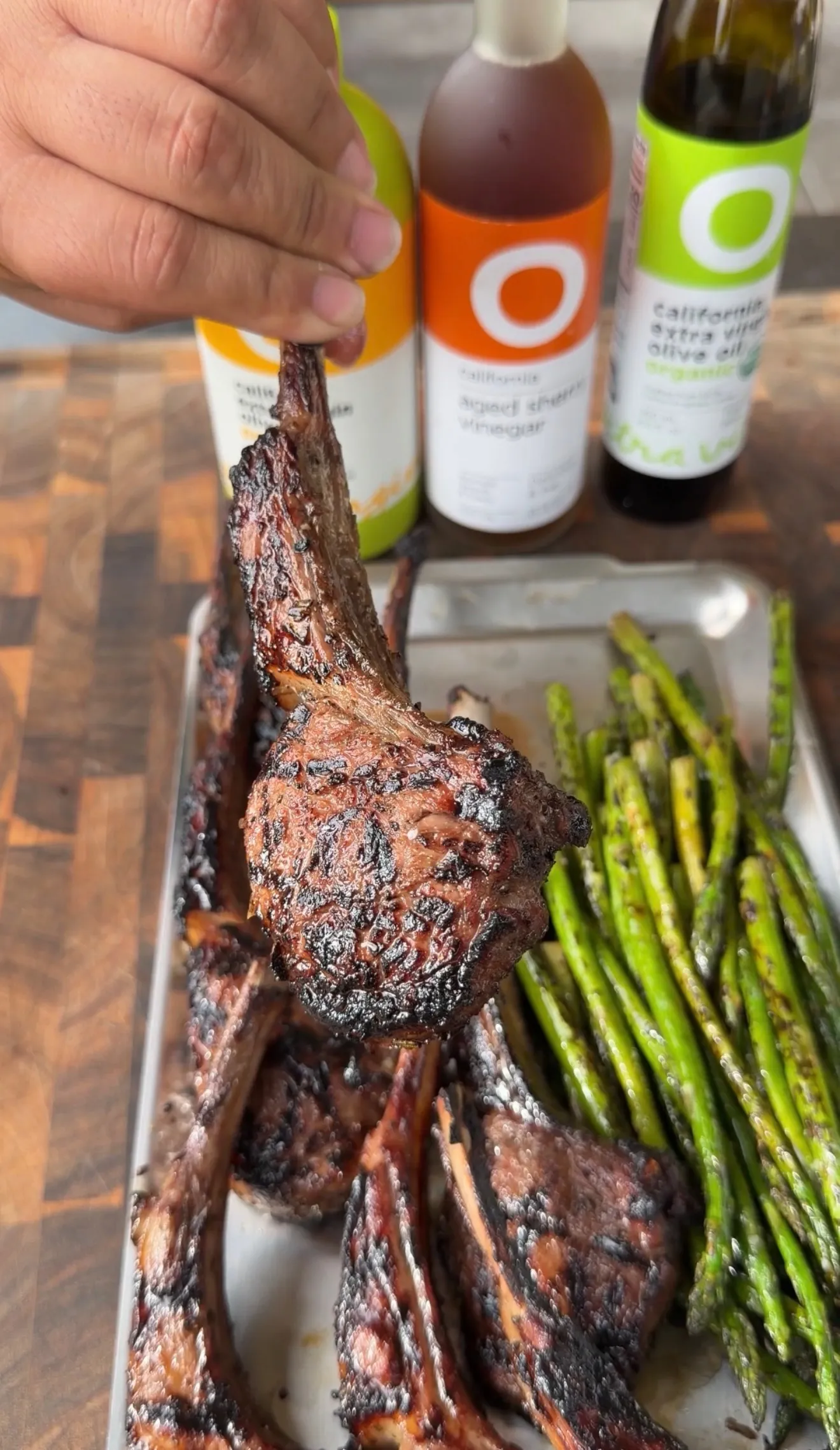 Hand holding a charred garlic sherry lamb lollipop chop over a tray of grilled lamb ribs and roasted asparagus