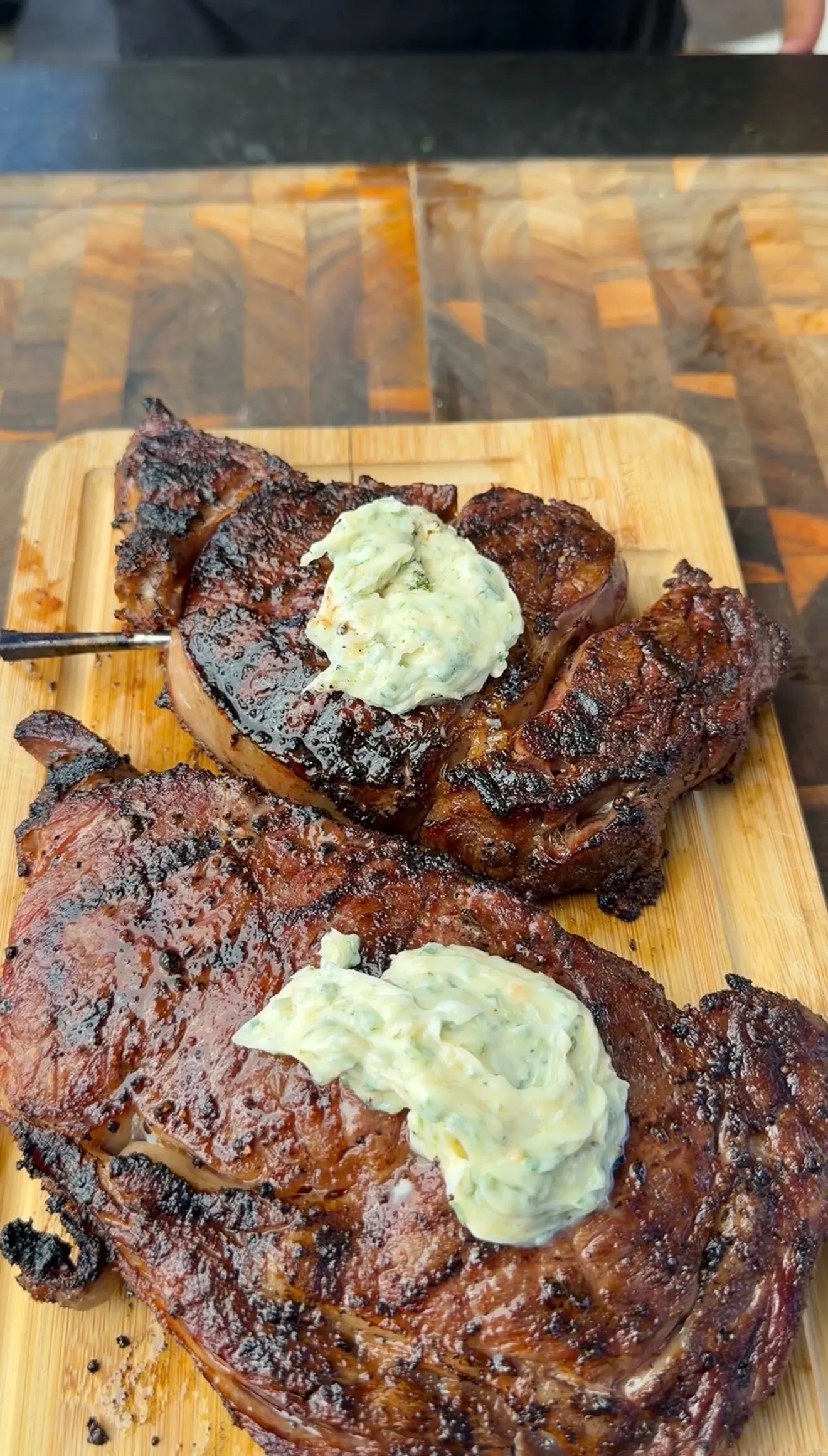 Two grilled ribeye steaks topped with melting herb butter on a wooden cutting board