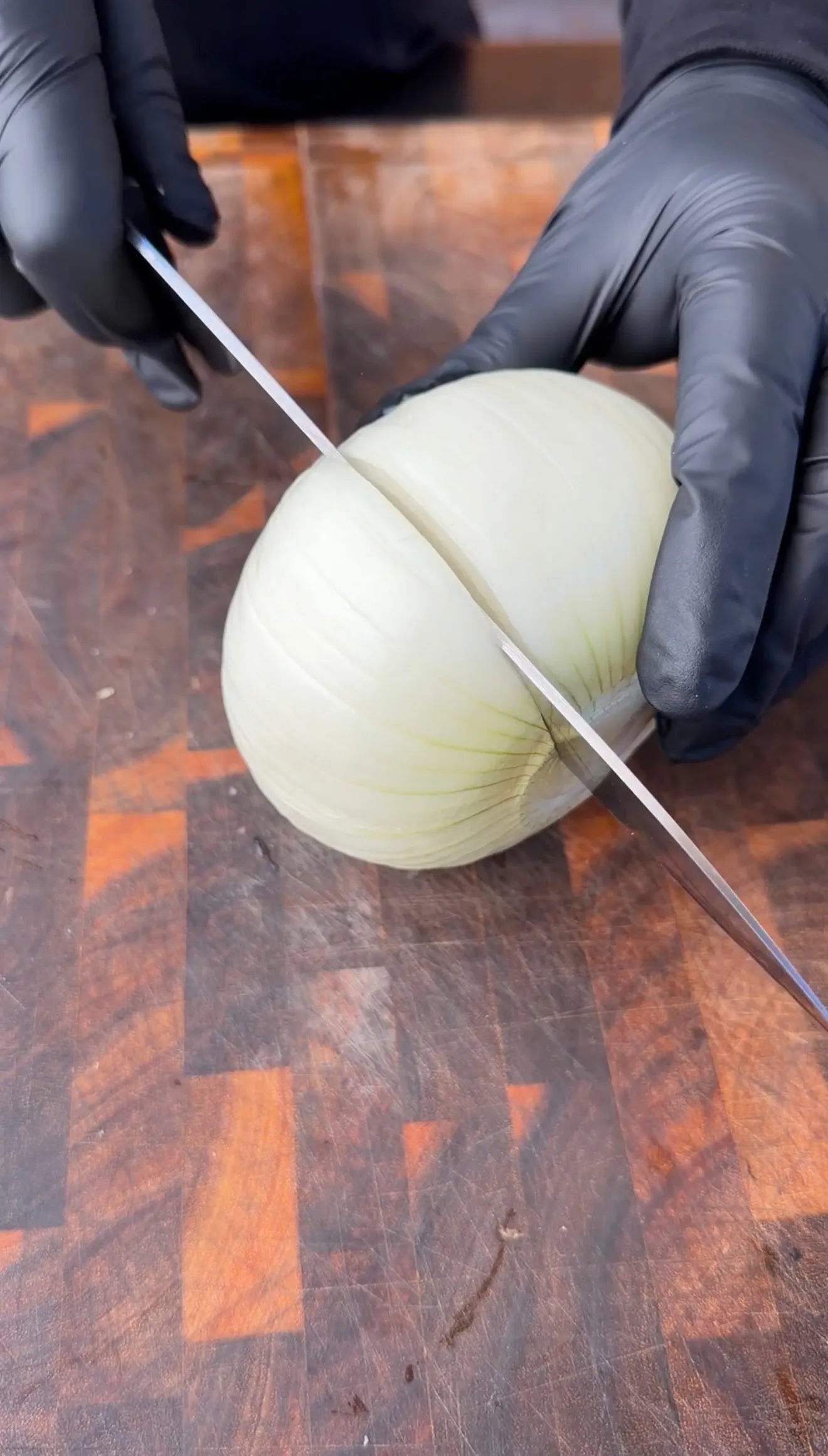 Gloved hands cutting a peeled white onion in half on a wooden cutting board with a sharp knife
