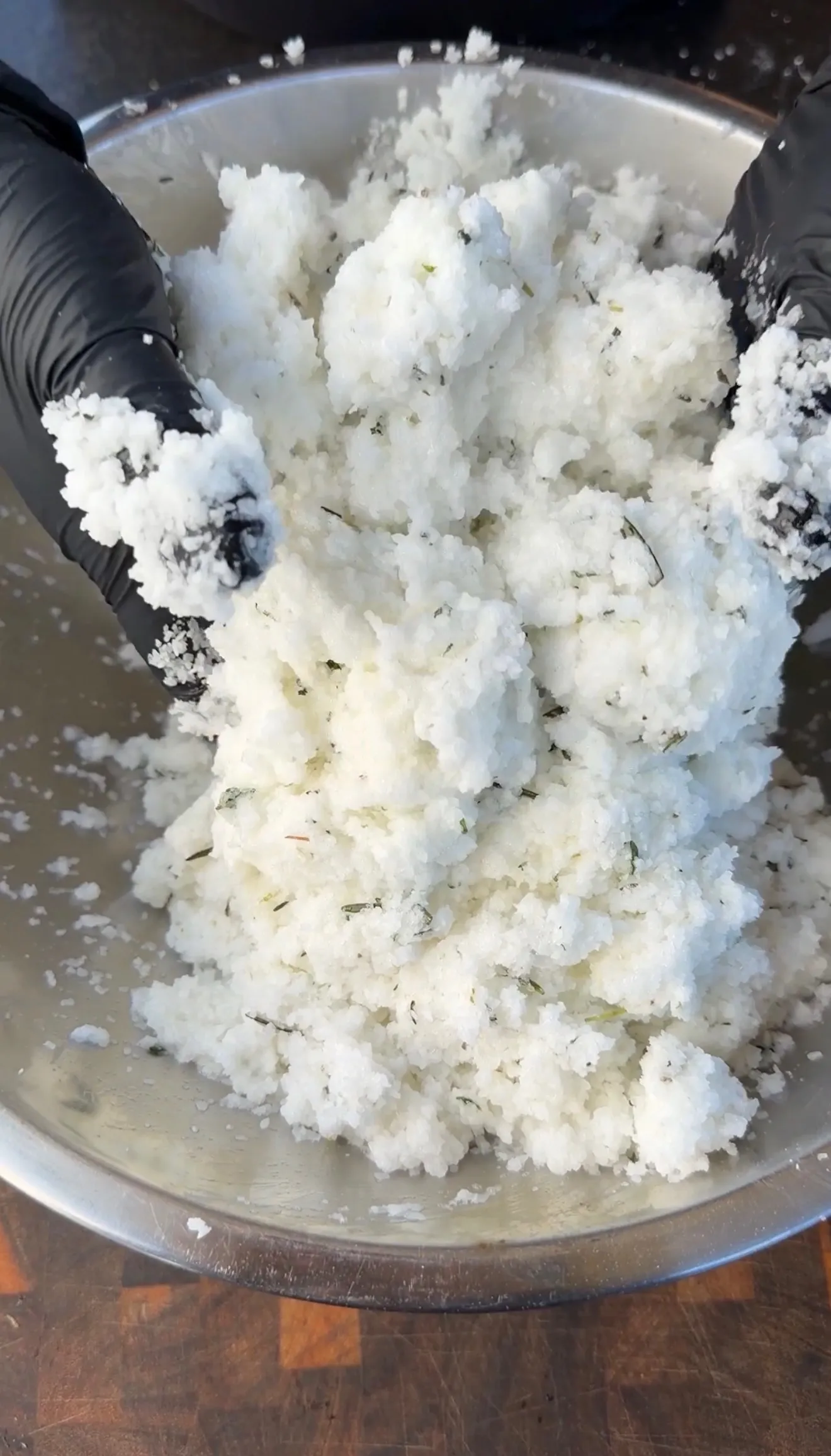 Hands in black gloves mixing coarse white salt with fresh herbs in a stainless steel bowl