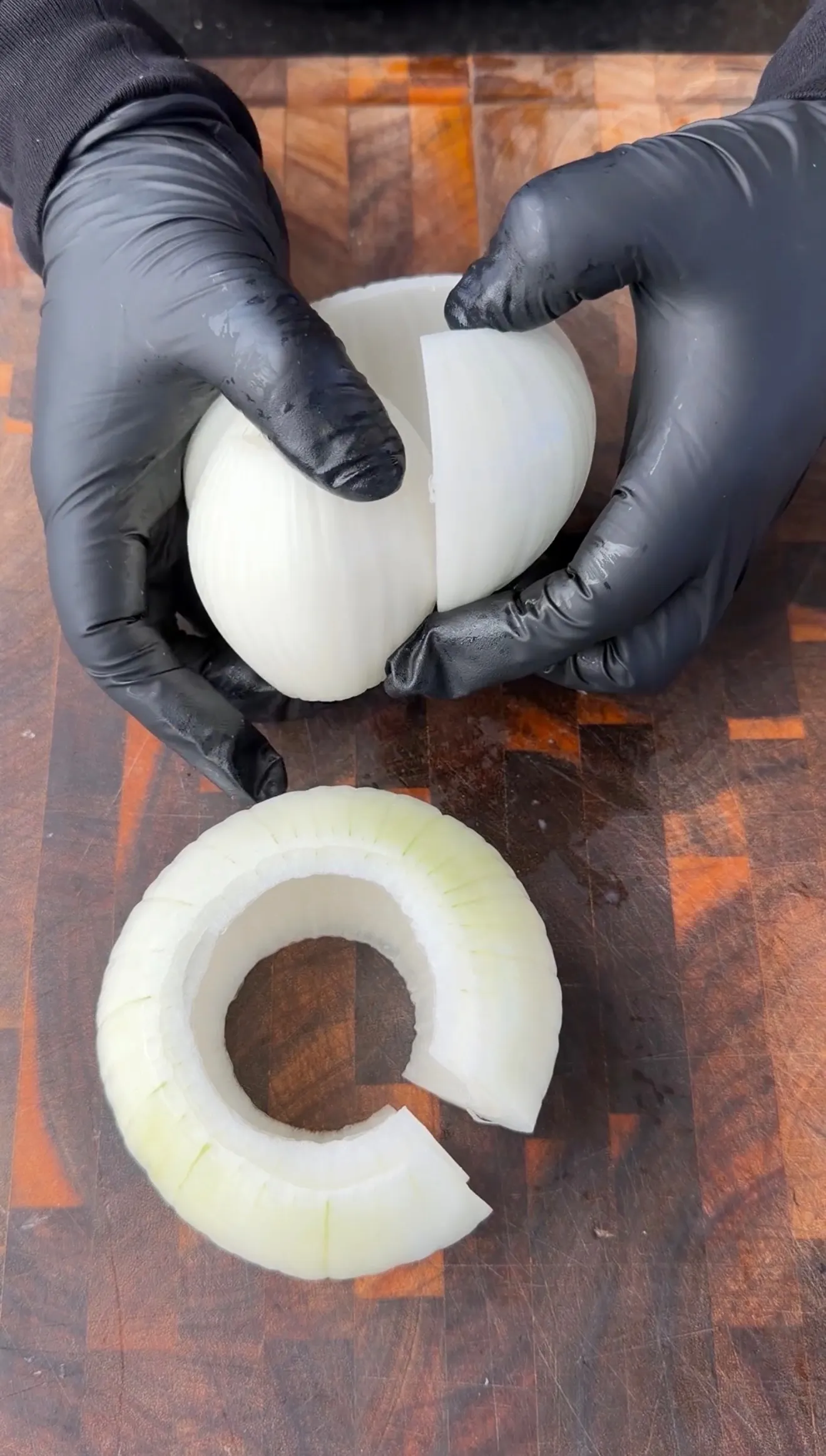Gloved hands holding a halved white onion above a scored hollowed onion ring on a wooden cutting board