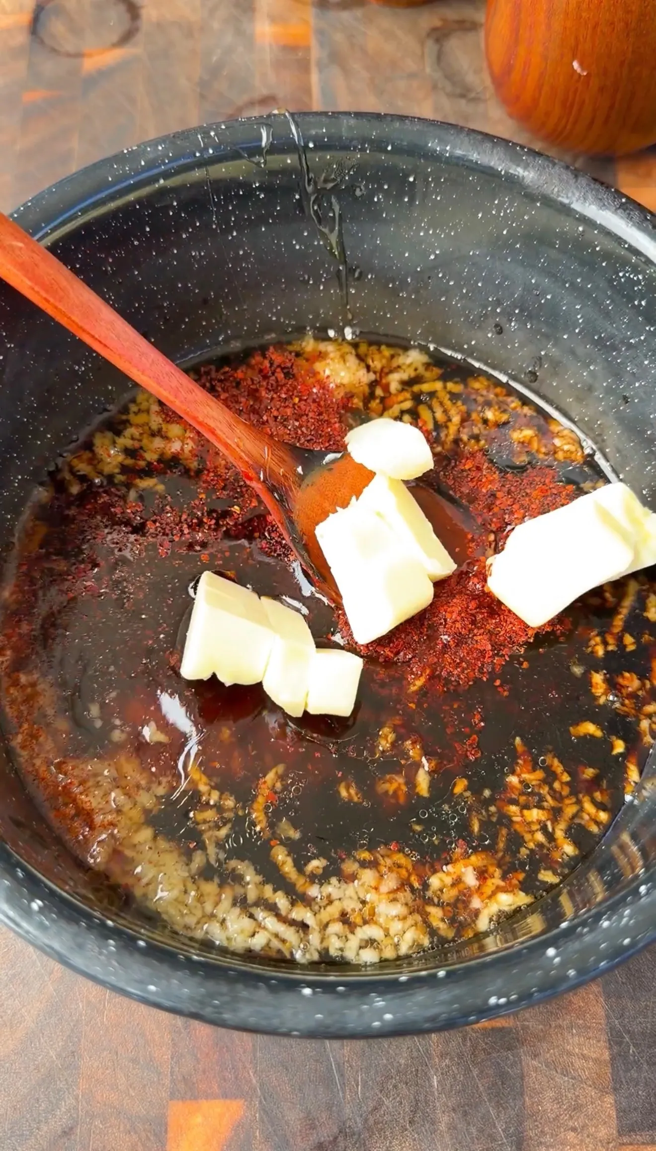 Butter pats and red chili powder added to honey garlic sauce in a dark saucepan with wooden spoon