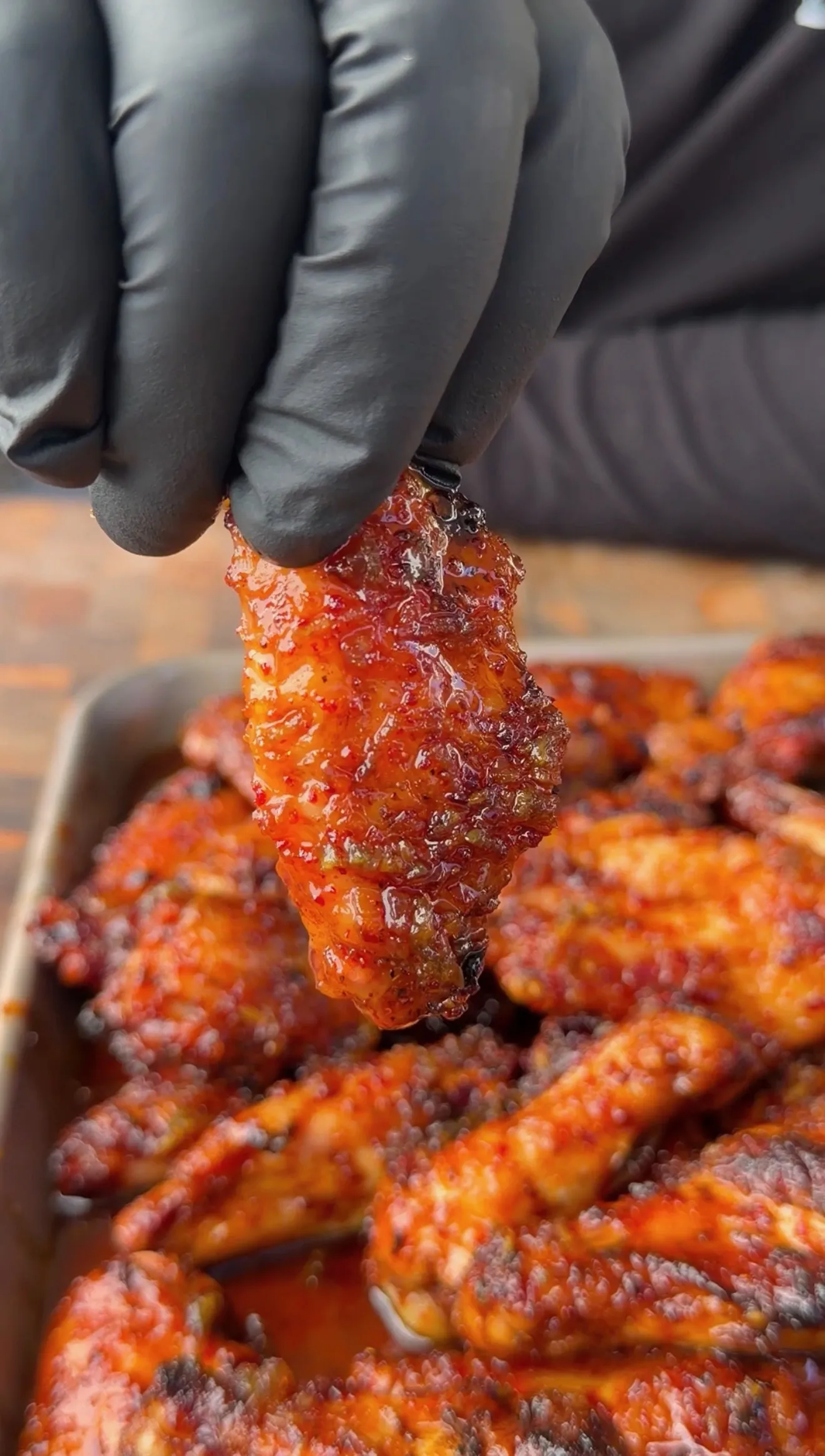 Glazed honey garlic chicken wings in a tray, one held up with black gloves showing sticky red sauce