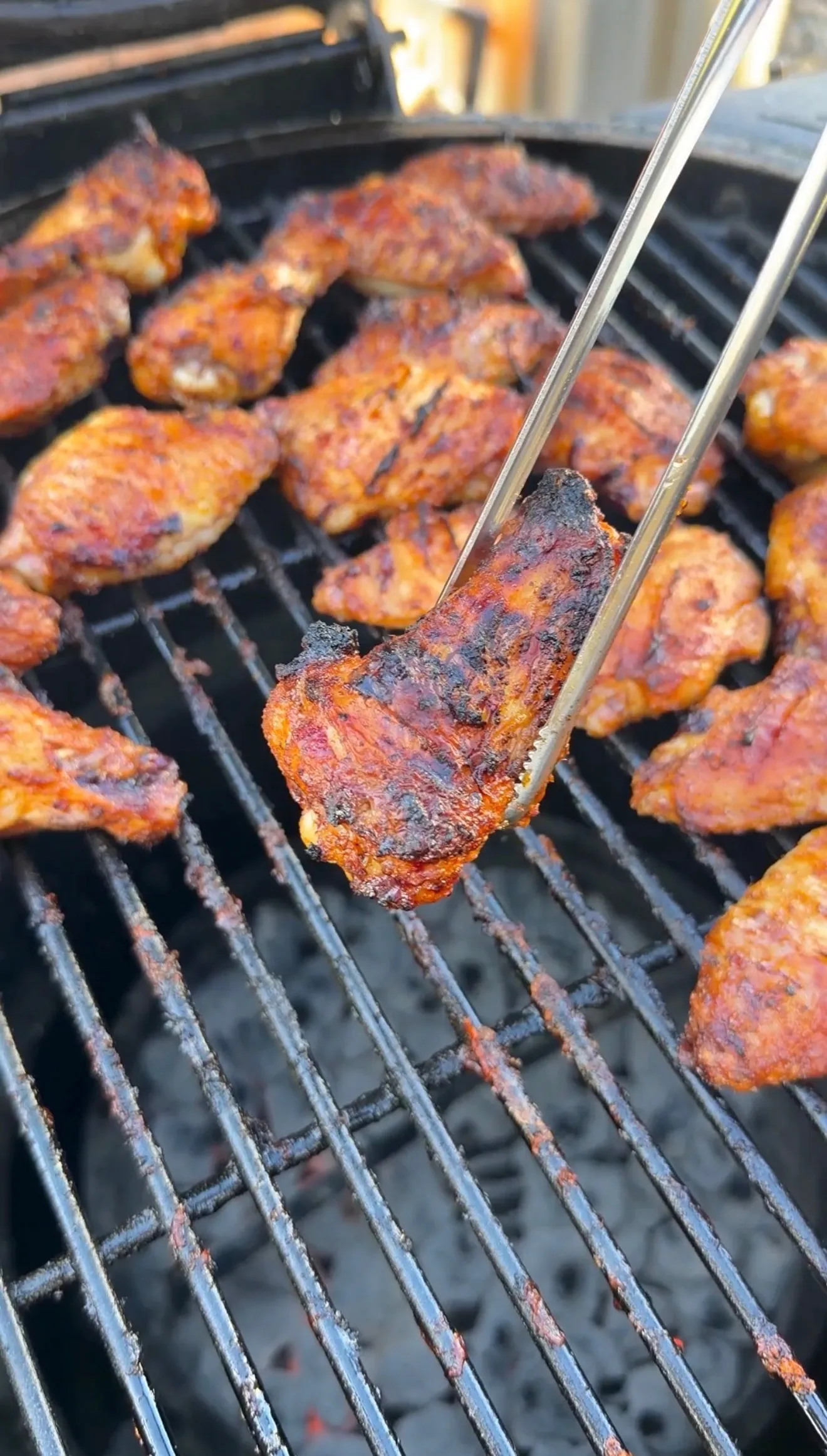 Honey garlic chicken wings grilling on charcoal grill with tongs holding one crispy caramelized wing