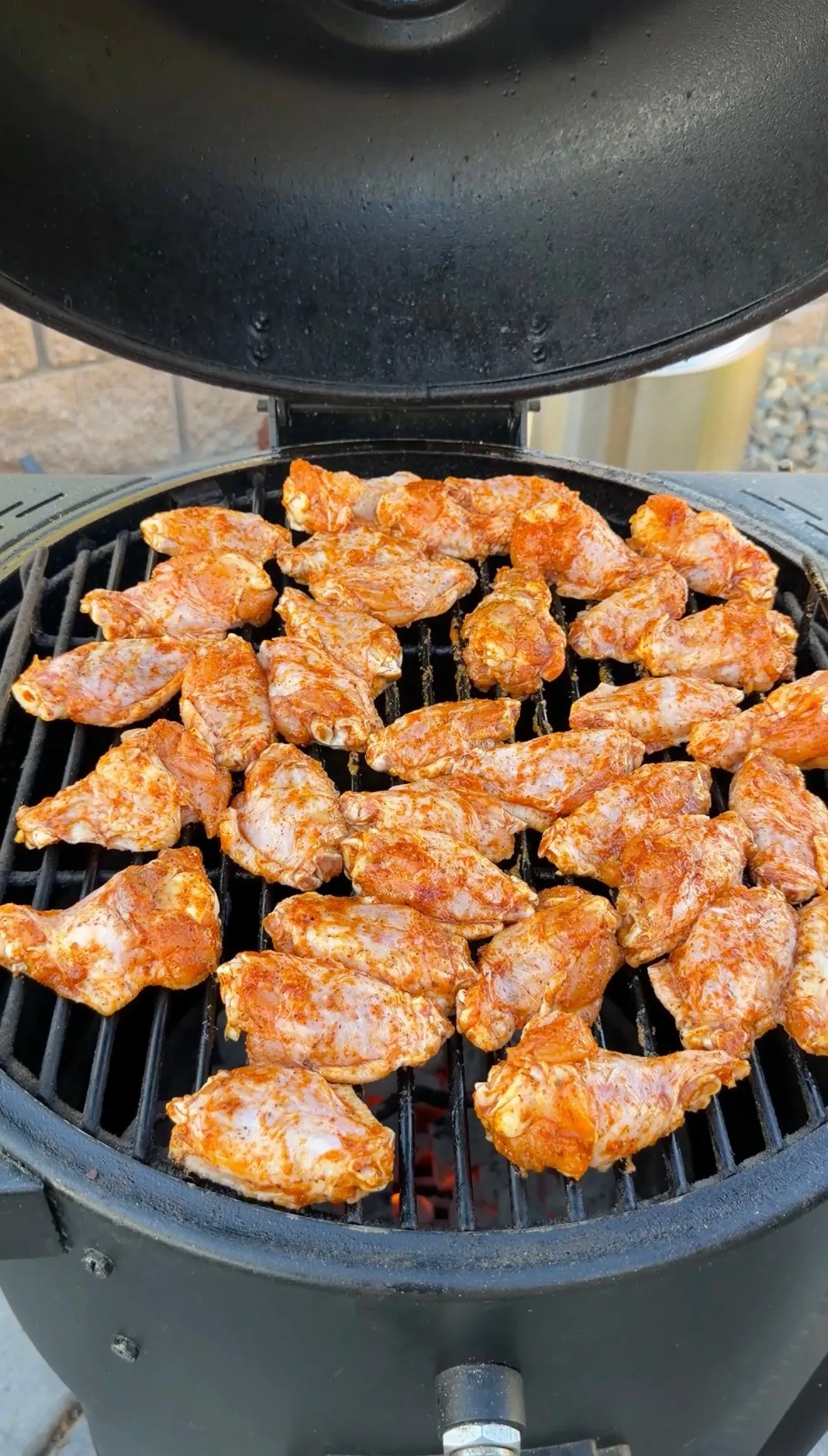 Seasoned honey garlic chicken wings arranged on a round charcoal kamado grill, coated in red spice rub