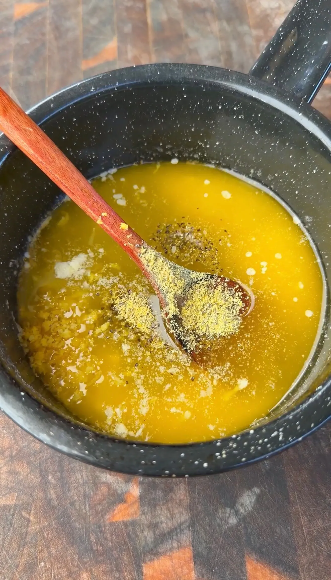 Golden honey lemon pepper butter sauce with seasonings in a dark speckled saucepan with wooden spoon