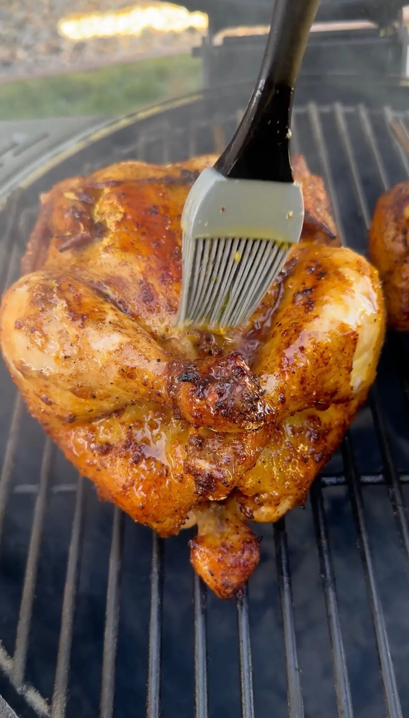 Whole spatchcock chicken on grill grates being basted with honey lemon pepper glaze using a silicone brush