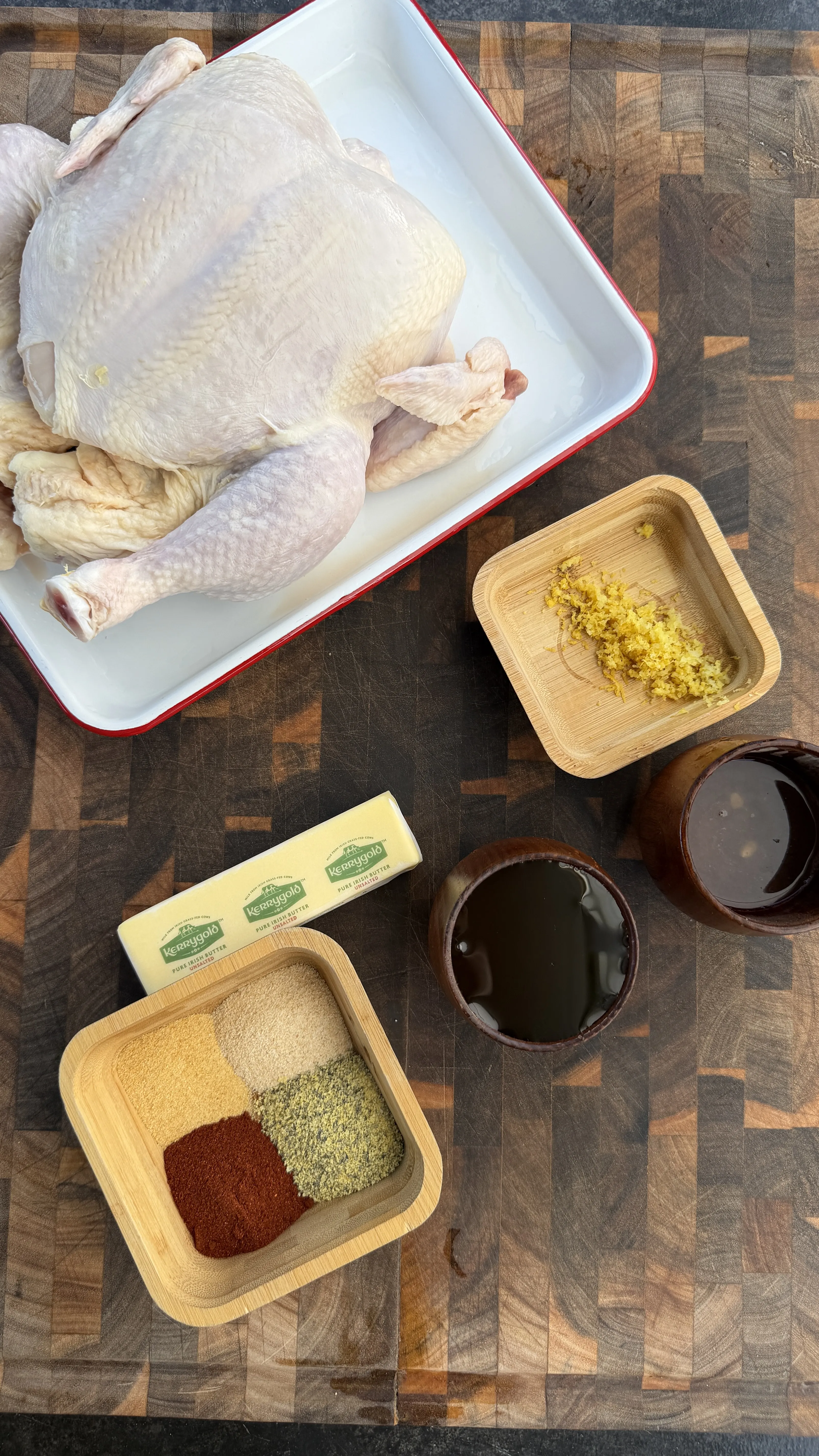 Raw whole chicken in a roasting pan with butter, spices, lemon zest, and honey on a wood cutting board