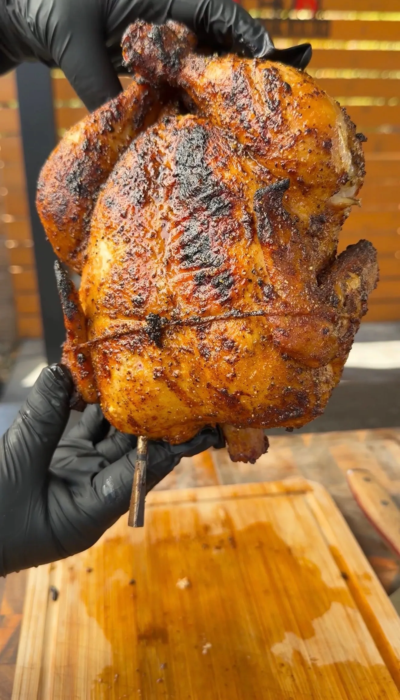 Hands in black gloves holding a golden whole rotisserie chicken with crispy spiced skin over a wooden cutting board