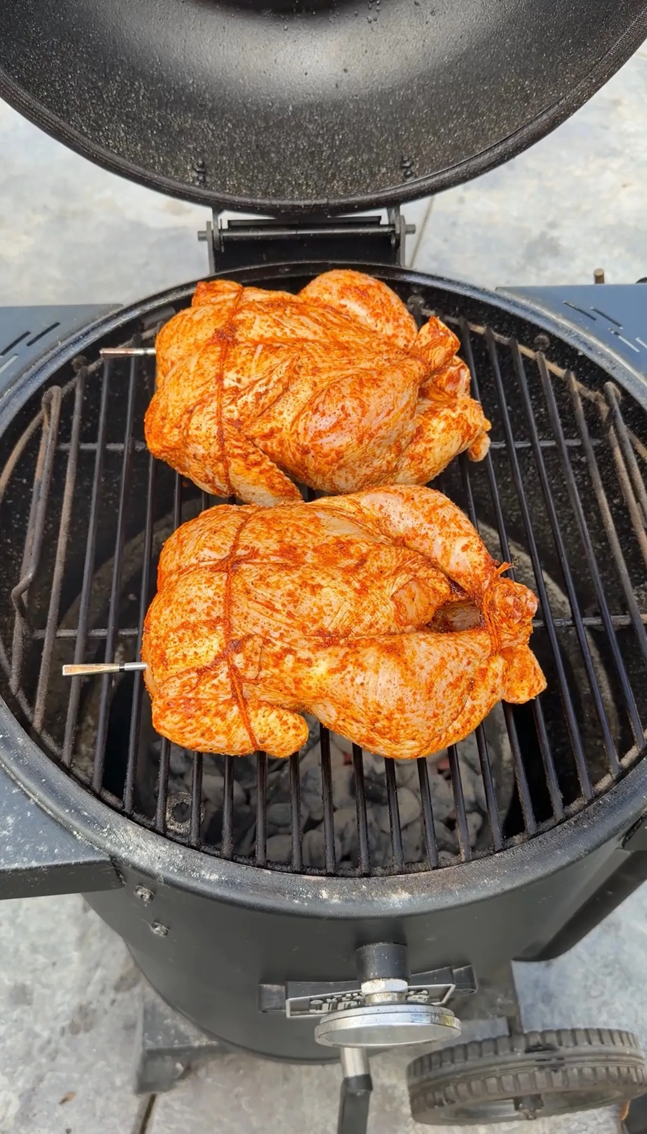 Two seasoned whole chickens trussed with twine, coated in spiced rub, placed on a round charcoal kettle grill