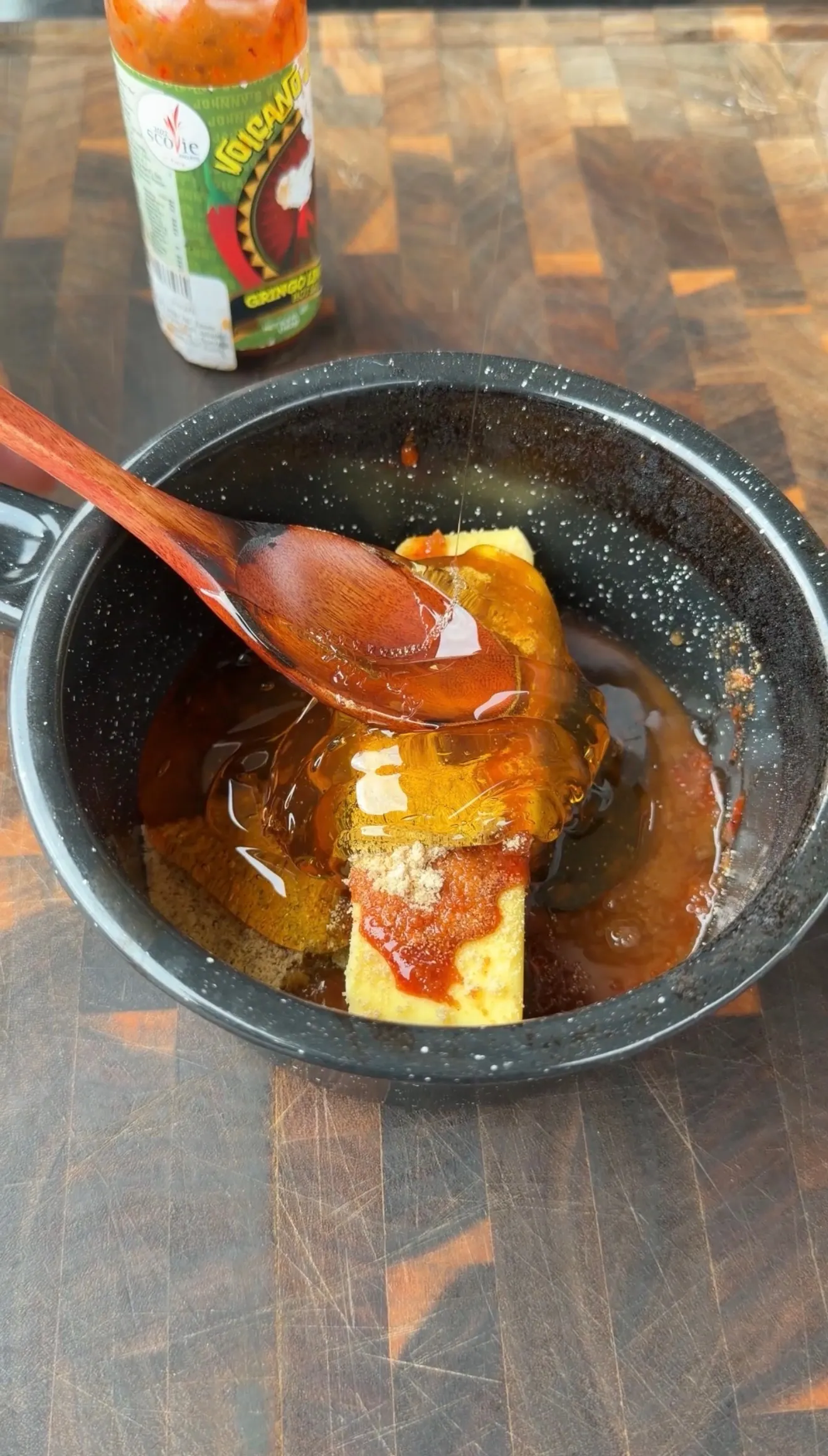 Spicy hot sauce glaze being spooned over ingredients in a black pan with Volcano hot sauce bottle in background