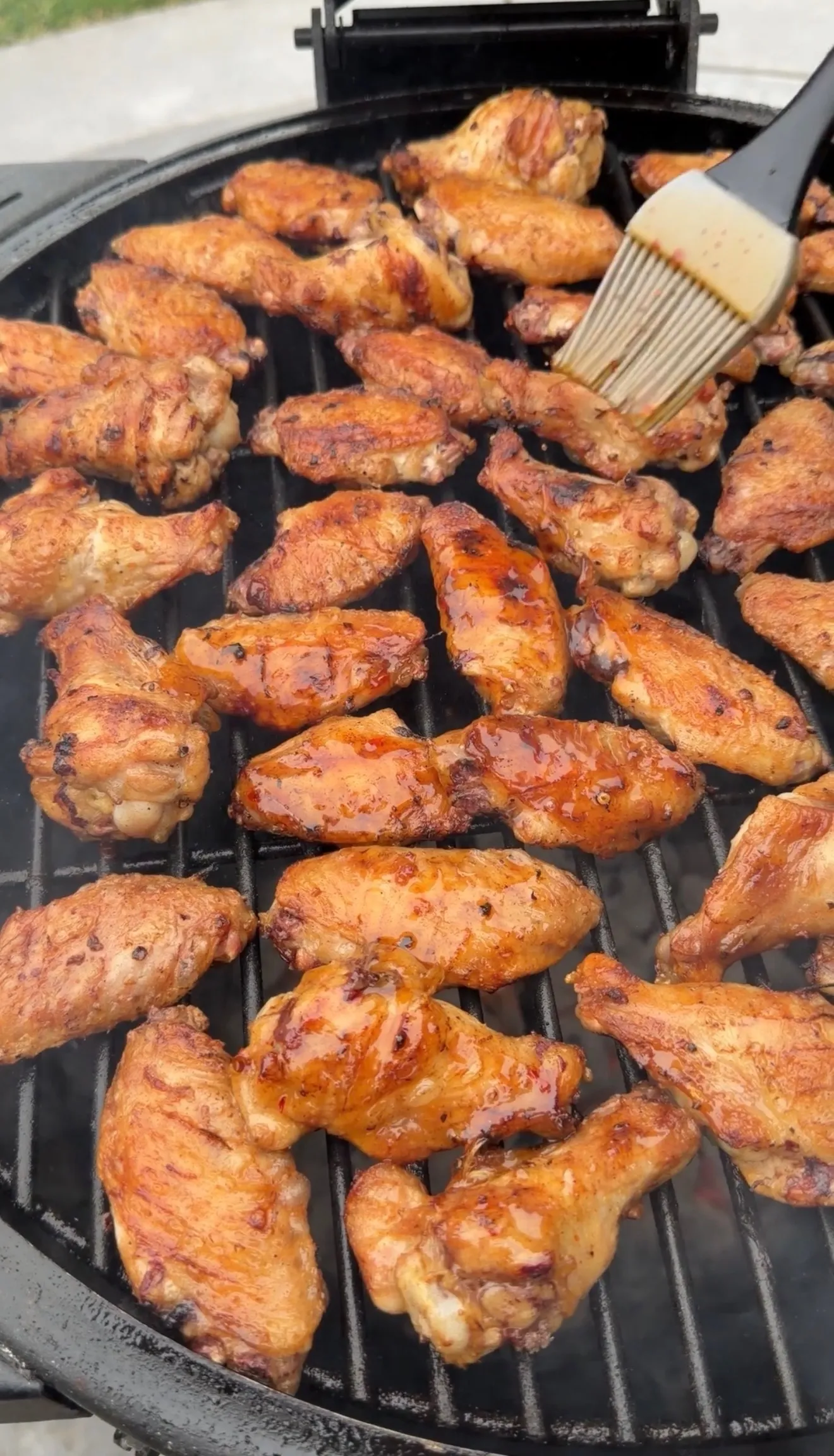 Dozens of glossy hot sauce glazed chicken wings on a round charcoal grill being basted with a pastry brush