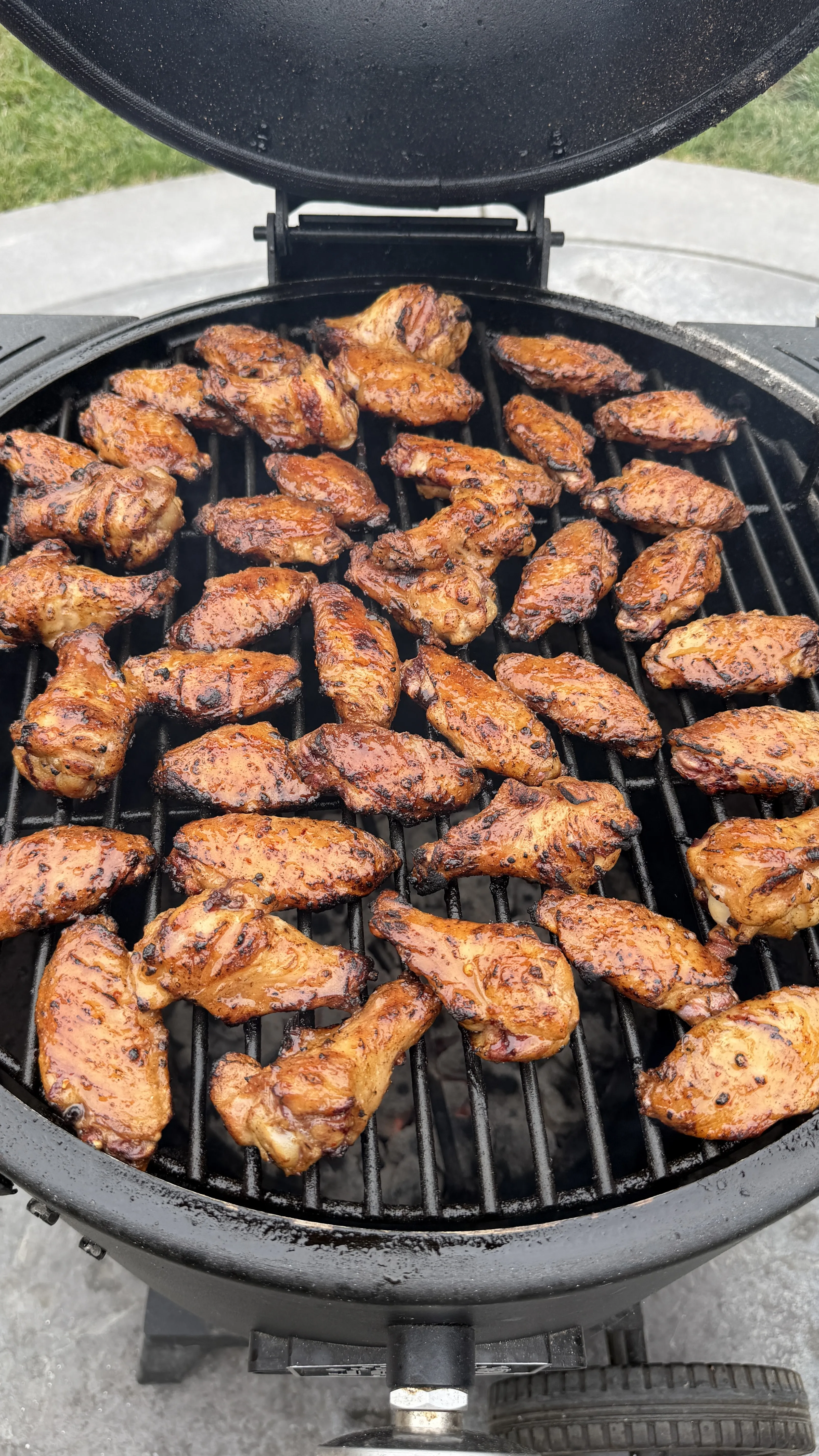 Dozens of hot sauce glazed chicken wings grilling on a round kamado charcoal grill with char marks