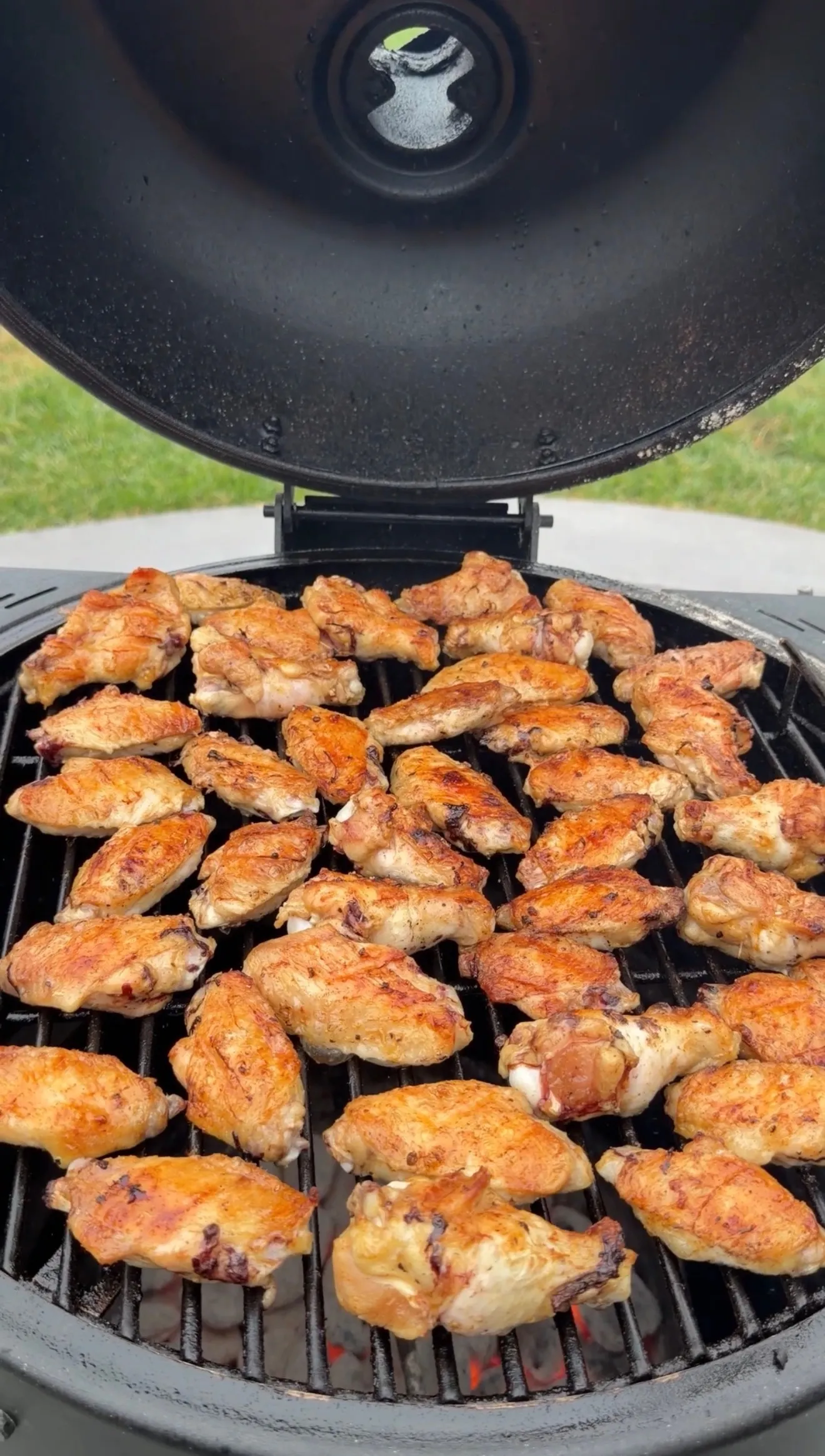 Dozens of golden hot sauce glazed chicken wings grilling on a round charcoal kamado grill outdoors