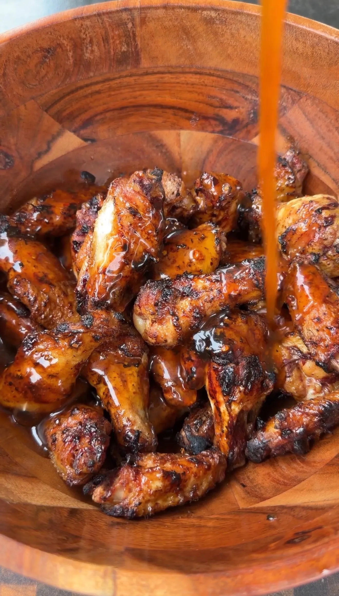 Grilled chicken wings being drizzled with glossy hot sauce in a rustic wooden bowl