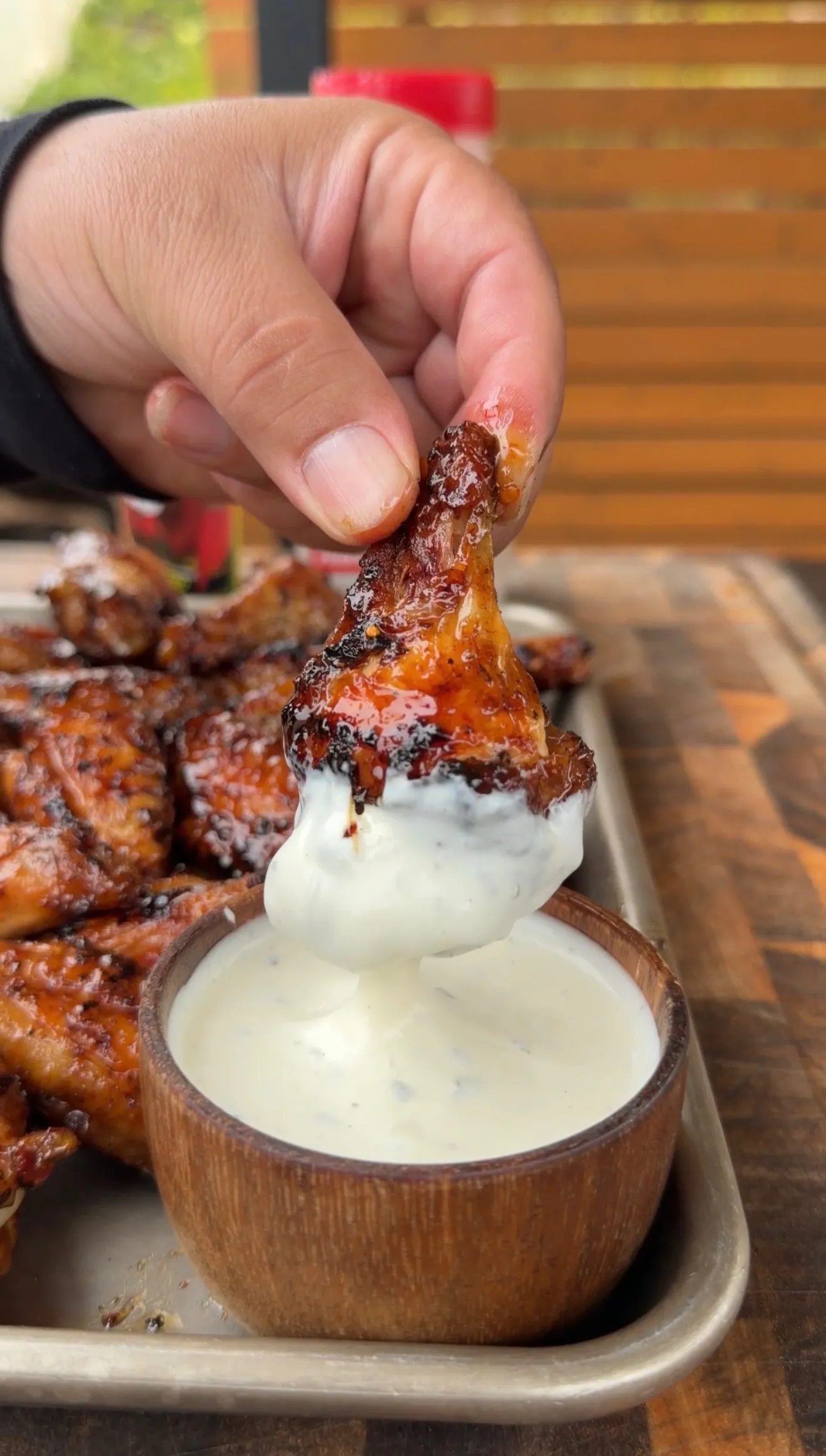 Hand dipping a glazed hot sauce chicken wing into a wooden bowl of creamy ranch dressing on a serving tray