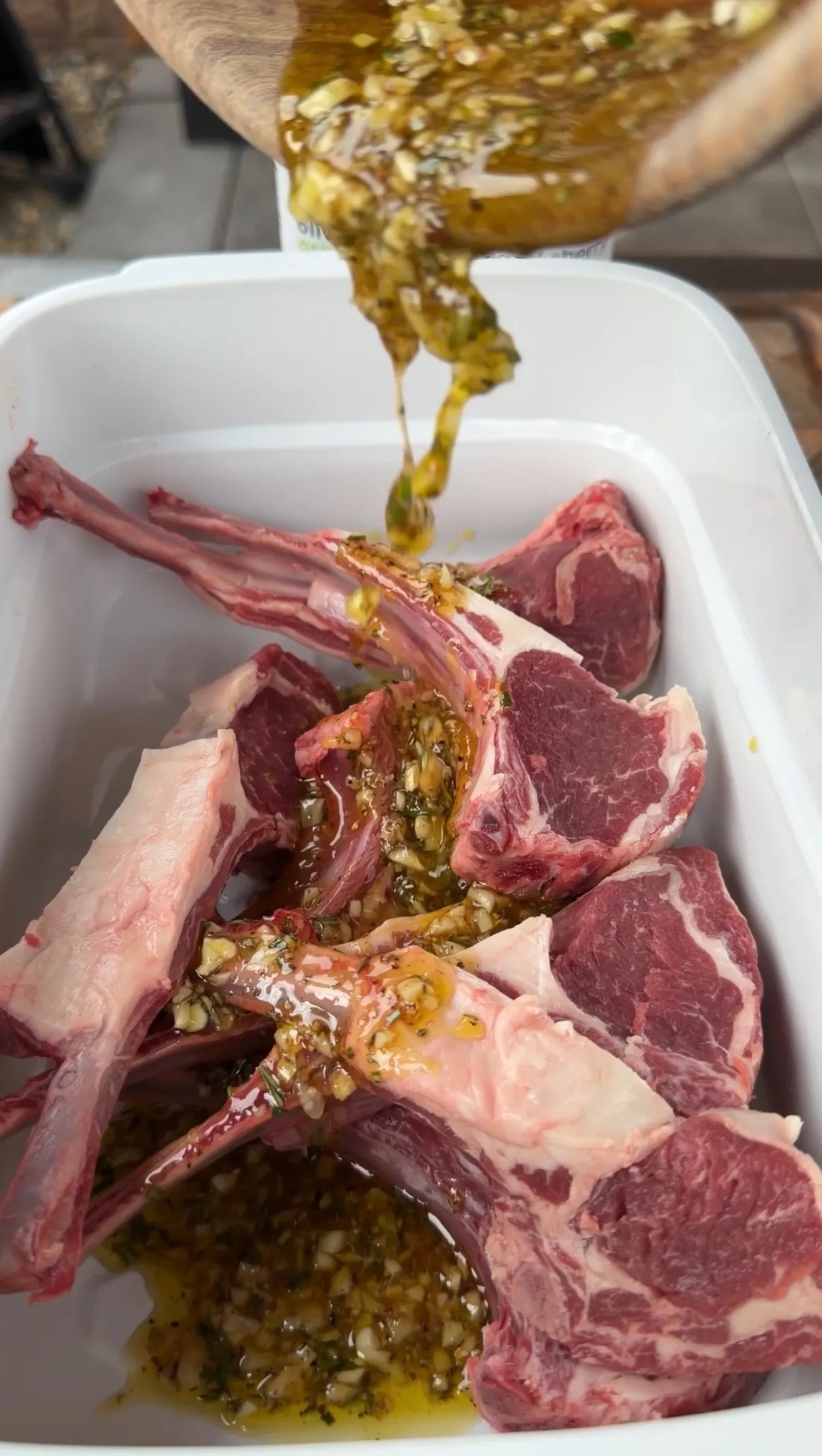 Raw lamb lollipop chops in a white container being coated with garlic, herb, and olive oil marinade