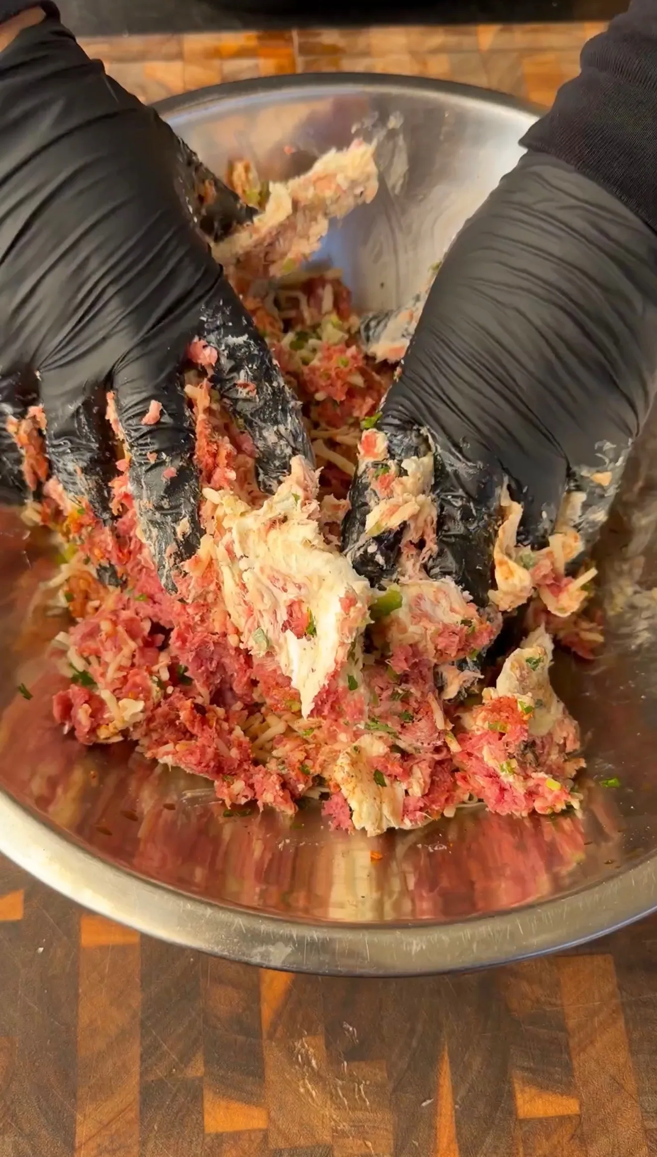 Hands in black gloves mixing seasoned ground beef mixture with herbs in a stainless steel bowl