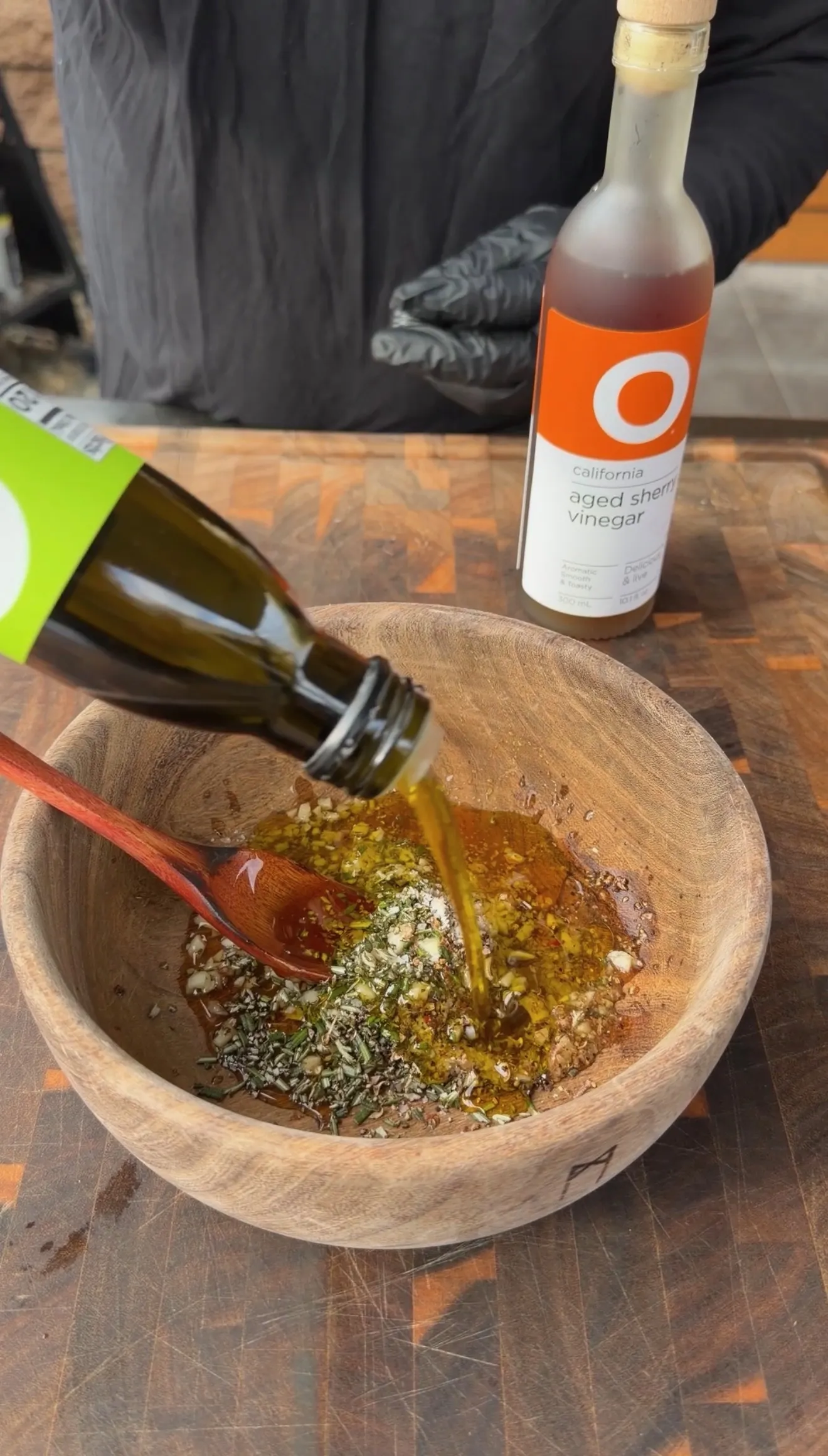 Olive oil being poured into a wooden bowl with herbs, garlic, and sherry vinegar for lamb lollipop marinade