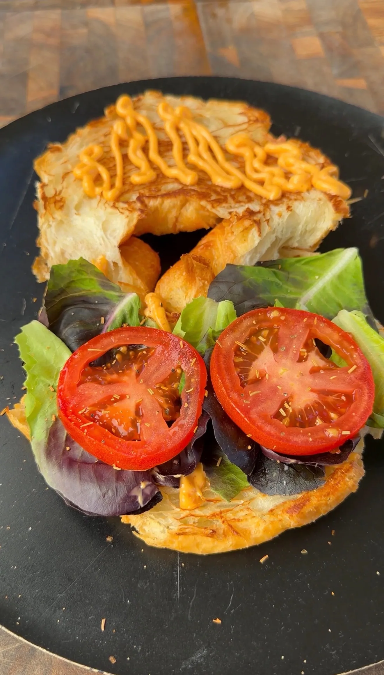 Open-face croissant burger with fresh tomato slices, mixed greens, and drizzled spicy sauce on a black plate