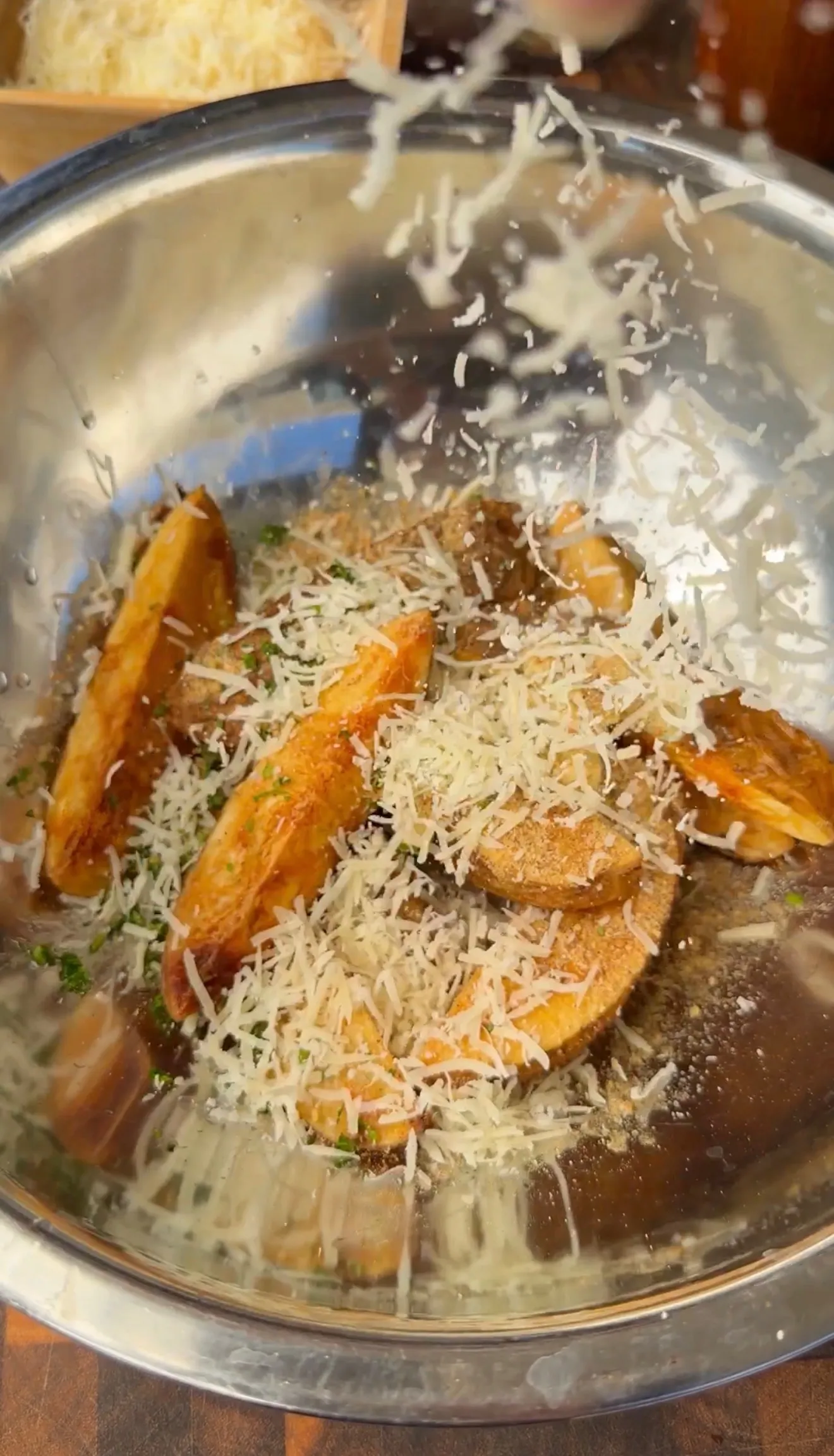 Crispy potato wedges being tossed with freshly grated Parmesan cheese and herbs in a stainless steel mixing bowl