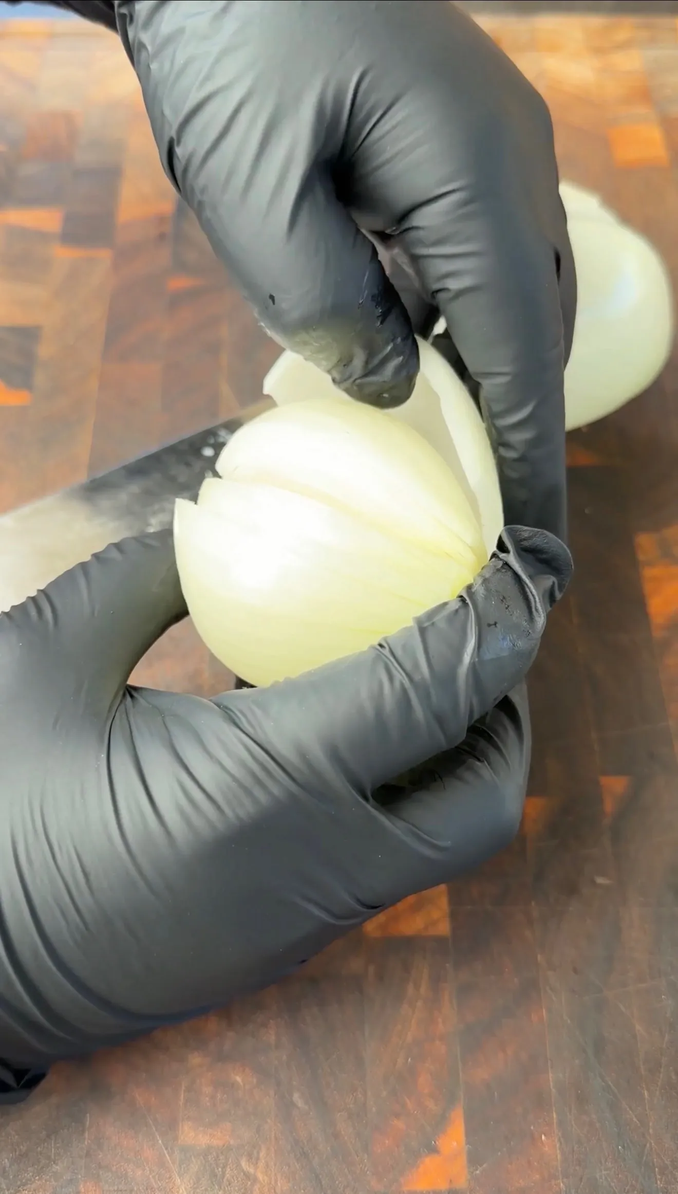Hands in black gloves peeling outer skin off a whole white onion on a wooden cutting board
