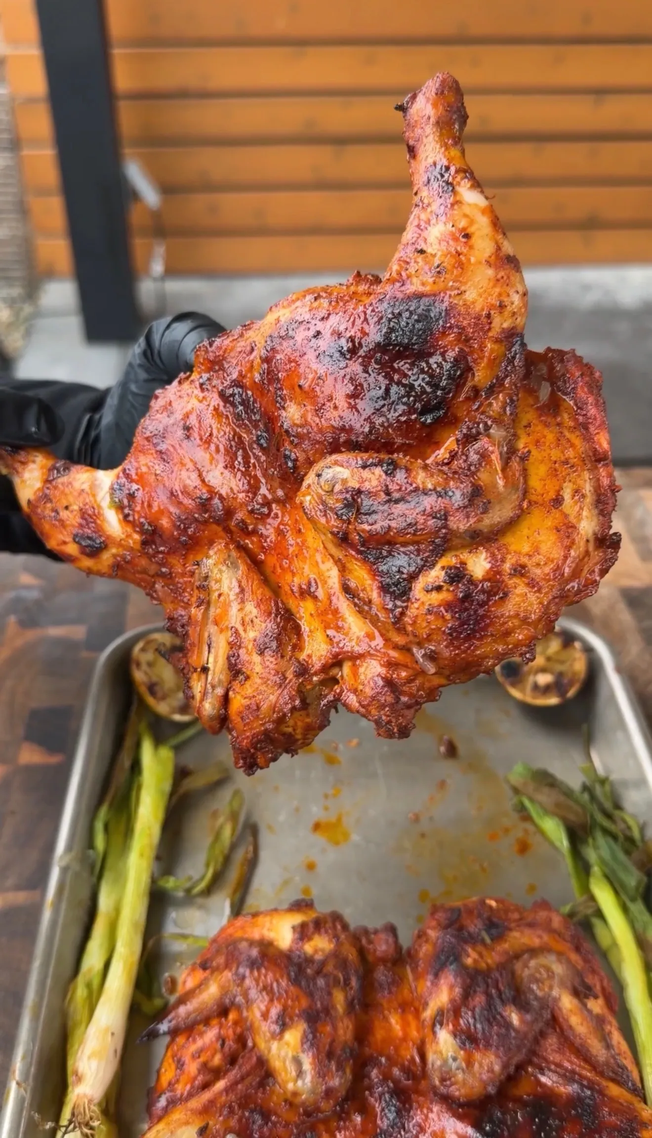 Charred and glazed Pollo Asado style Cornish hen held up in black gloved hand over roasting pan with green onions